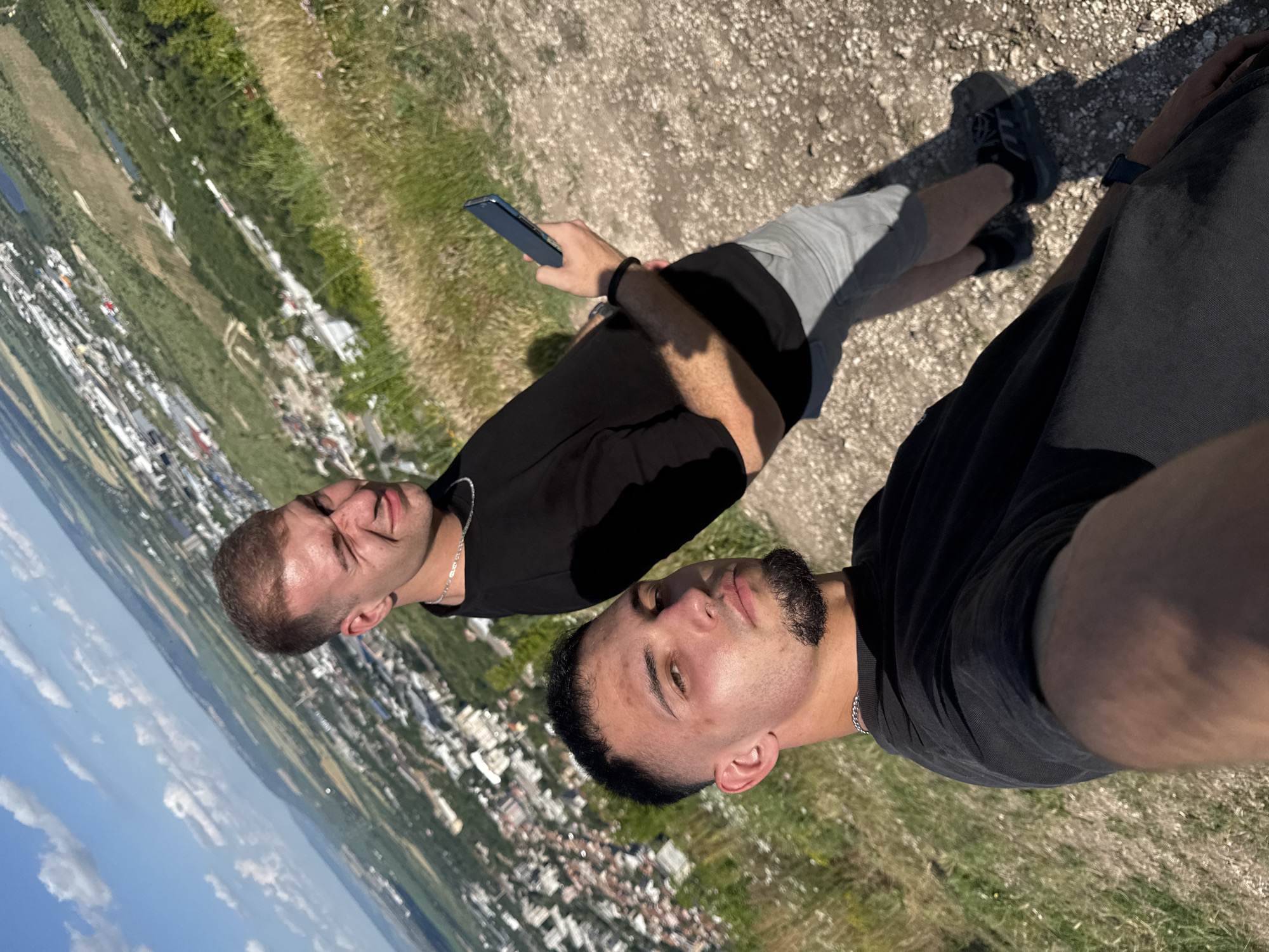 The image shows two young men taking a selfie on a hill overlooking the city of Shumen, Bulgaria. The man in the foreground is closer to the camera, wearing a black t-shirt and a silver chain. He has short dark hair and a goatee. The man in the background is further away, also wearing a black t-shirt and grey shorts. He is holding a smartphone in his right hand and winking at the camera. The city of Shumen is visible in the background, with buildings and greenery spread across the landscape. The sky is blue with some white clouds. The ground in the foreground is covered with gravel and dry grass. The scene appears to be set on a sunny day.