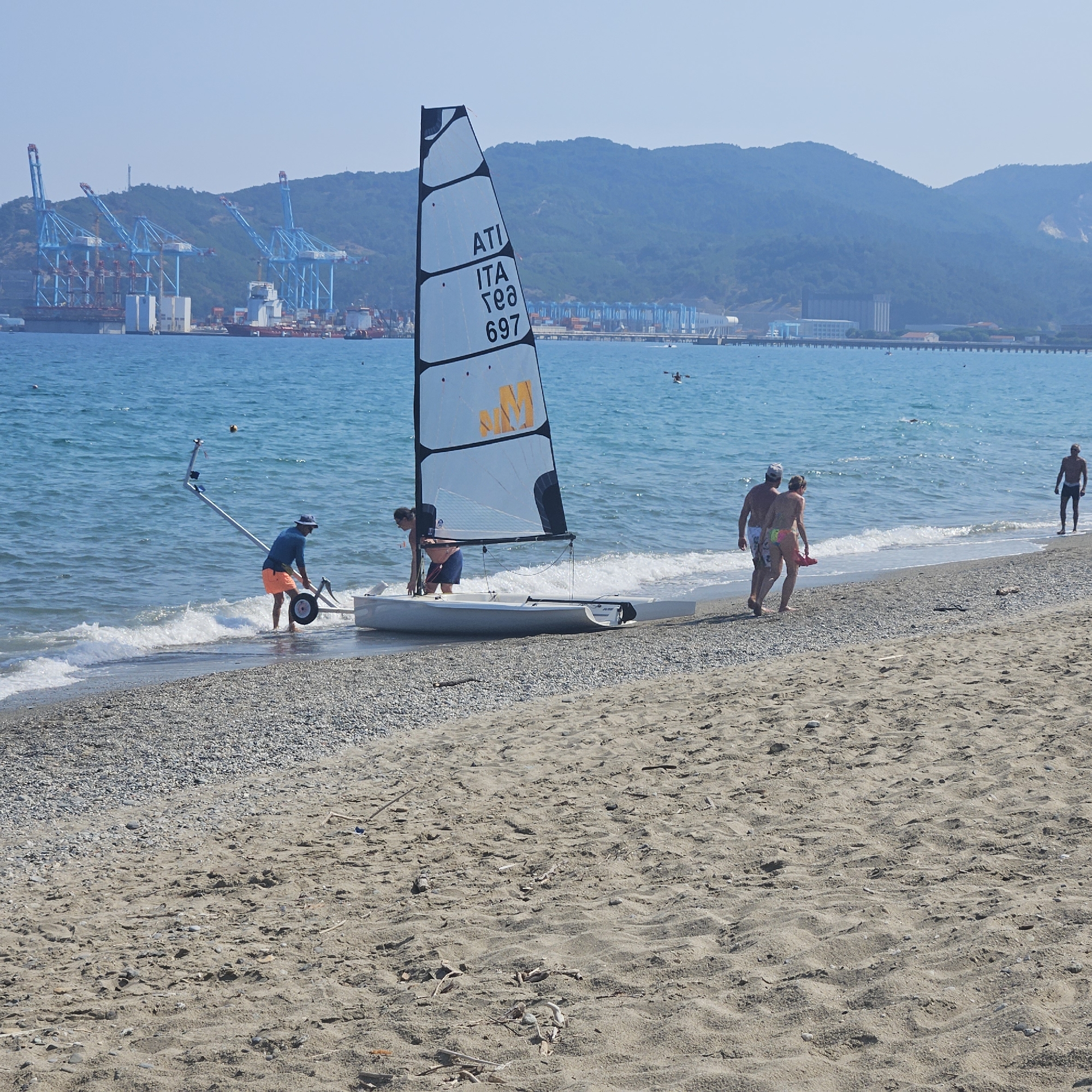 The image depicts a sunny day at a beach in Savona, Italy, with a small sailboat being prepared near the shore. In the foreground, the sand is covered with pebbles, and some driftwood is scattered around. A man in an orange shirt and blue shorts is at the water's edge, attending to a small sailboat that is partially submerged in the shallow water. The sailboat has a white sail with black trim and the letters "ATI ITA TEO 697" visible. Another person, partially visible from behind, is also near the sailboat. Further out in the turquoise sea, two people in swimwear are walking along the shoreline, and a lone figure stands on the wet sand. In the background, a large industrial port with multiple cranes is visible, along with a lush, green mountain range. The sky is clear and bright blue.