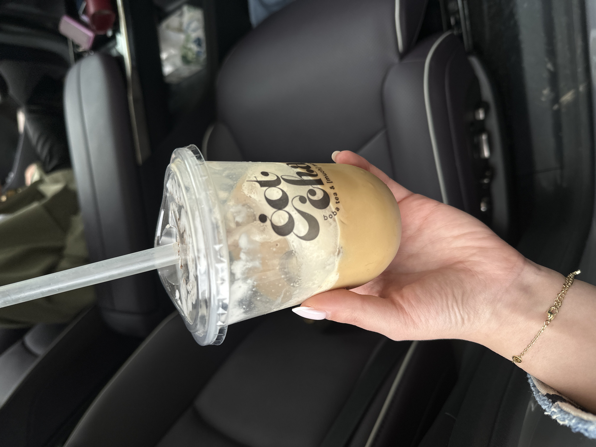 A hand is holding a partly consumed cup of boba tea inside a vehicle with dark leather interior. The cup has a clear plastic lid and a clear straw. The tea itself is light brown. The cup has a logo on it. The person wearing the bracelet, is inside a car with dark leather seats, it is impossible to tell the location in the city of Yerevan, Armenia.