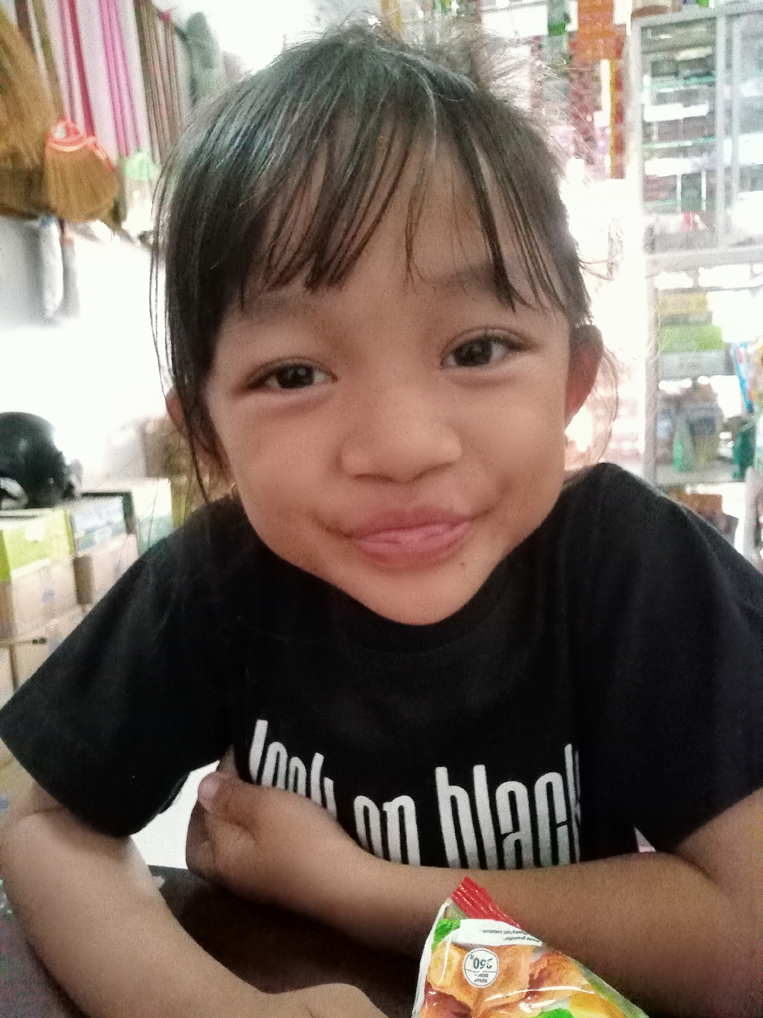 A young Asian girl with dark hair is sitting at a table, looking at the camera with a slight smile and her tongue peeking out. She is wearing a black t-shirt with white text that reads "look on black". Her arms are resting on the table, and a bag of snacks is visible in the foreground. The background is blurred but appears to be a store or market, with shelves of goods and a broom visible on the left side. The image was taken in Simo, Indonesia.