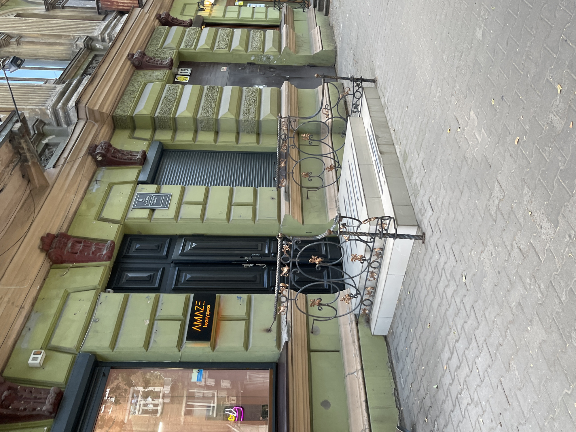 The media file depicts a street view of a building facade in Dnipro, Ukraine. The building is painted a pale green and features ornate, dark brown decorative elements around its entrances and windows. The facade is characterized by several recessed doorways and windows, with a metal security gate visible over one of the doorways. A wrought-iron railing with decorative gold accents adorns a raised platform in front of one of the entrances. The walkway in front of the building is paved with interlocking grey bricks, and scattered leaves suggest it might be autumn. A sign with the text "AMAZ E beauty space" in orange and white lettering is prominently displayed next to an entrance. The overall impression is of an urban setting with a touch of historical architecture. The lighting suggests it is daytime, and the weather appears to be clear. There are no people visible in the frame.