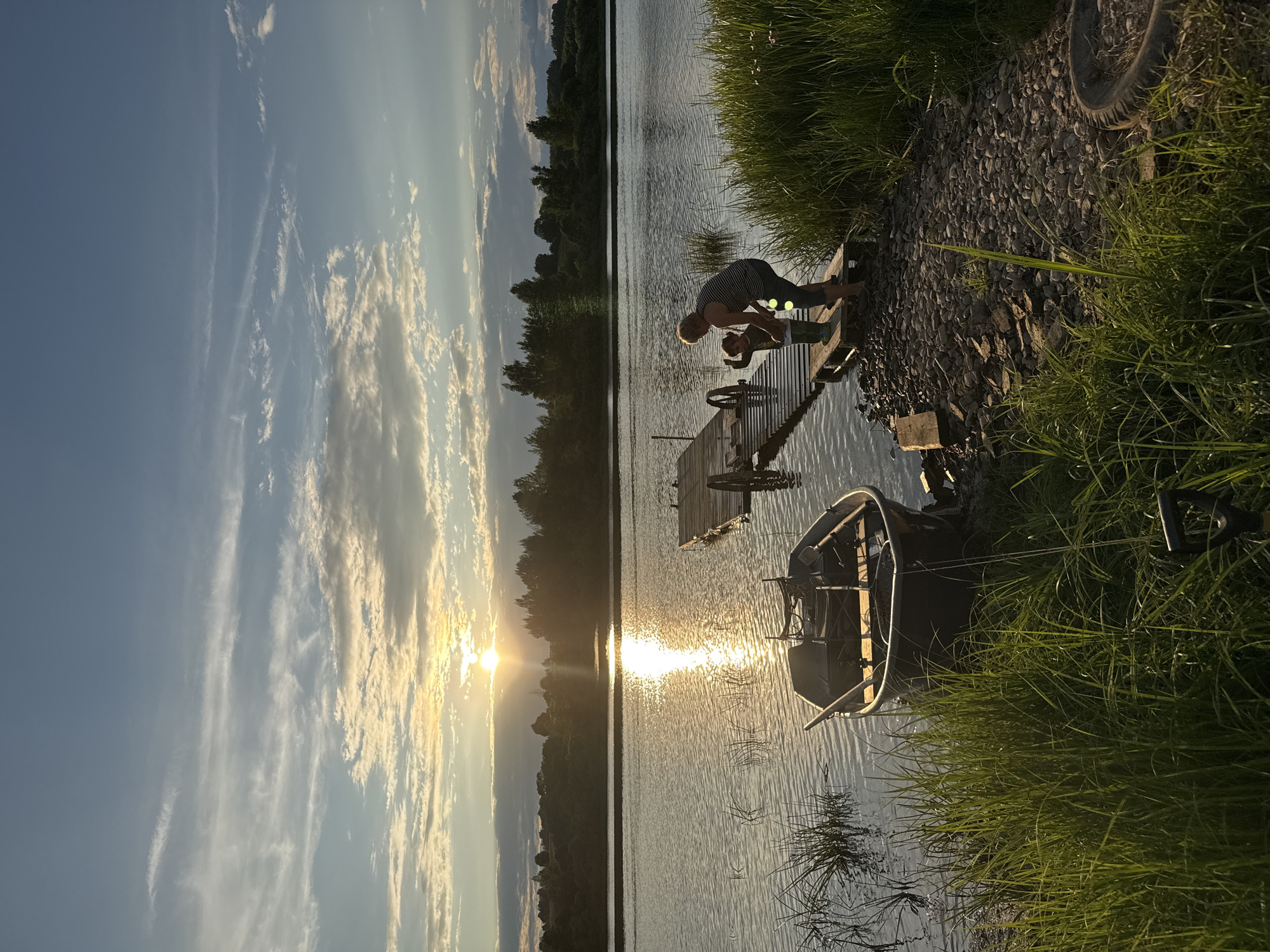 This image depicts a sunset scene at a lake, possibly located near Sitno, Russia. In the foreground, a dark-colored boat is tied to the grassy shore. A wooden dock extends into the water, with two figures, an adult and a child, standing on it. The adult, wearing a striped shirt, appears to be interacting with the child. The lake reflects the warm glow of the setting sun, which is visible in the distance behind a line of trees. The sky is filled with scattered clouds, illuminated by the sun. The overall atmosphere is serene and peaceful, characteristic of a rural or natural setting.