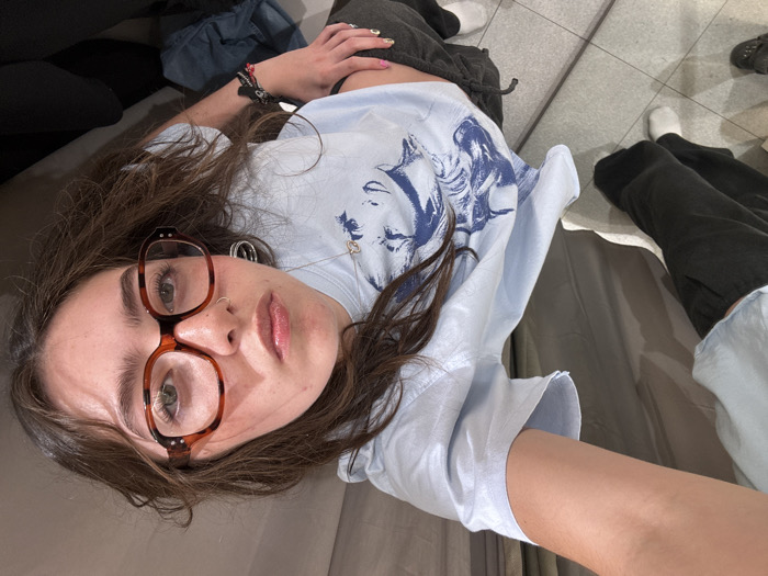 A woman lies on a beige surface, likely in a fitting room, judging by the curtains. She wears glasses, a blue t-shirt with a graphic, and dark sweatpants. She also wears a silver necklace and has multiple piercings in her ears and nose. Parts of her body and clothes are visible around her. The background shows a tiled floor, clothing items, and presumably her feet. The perspective suggests she is taking a selfie.