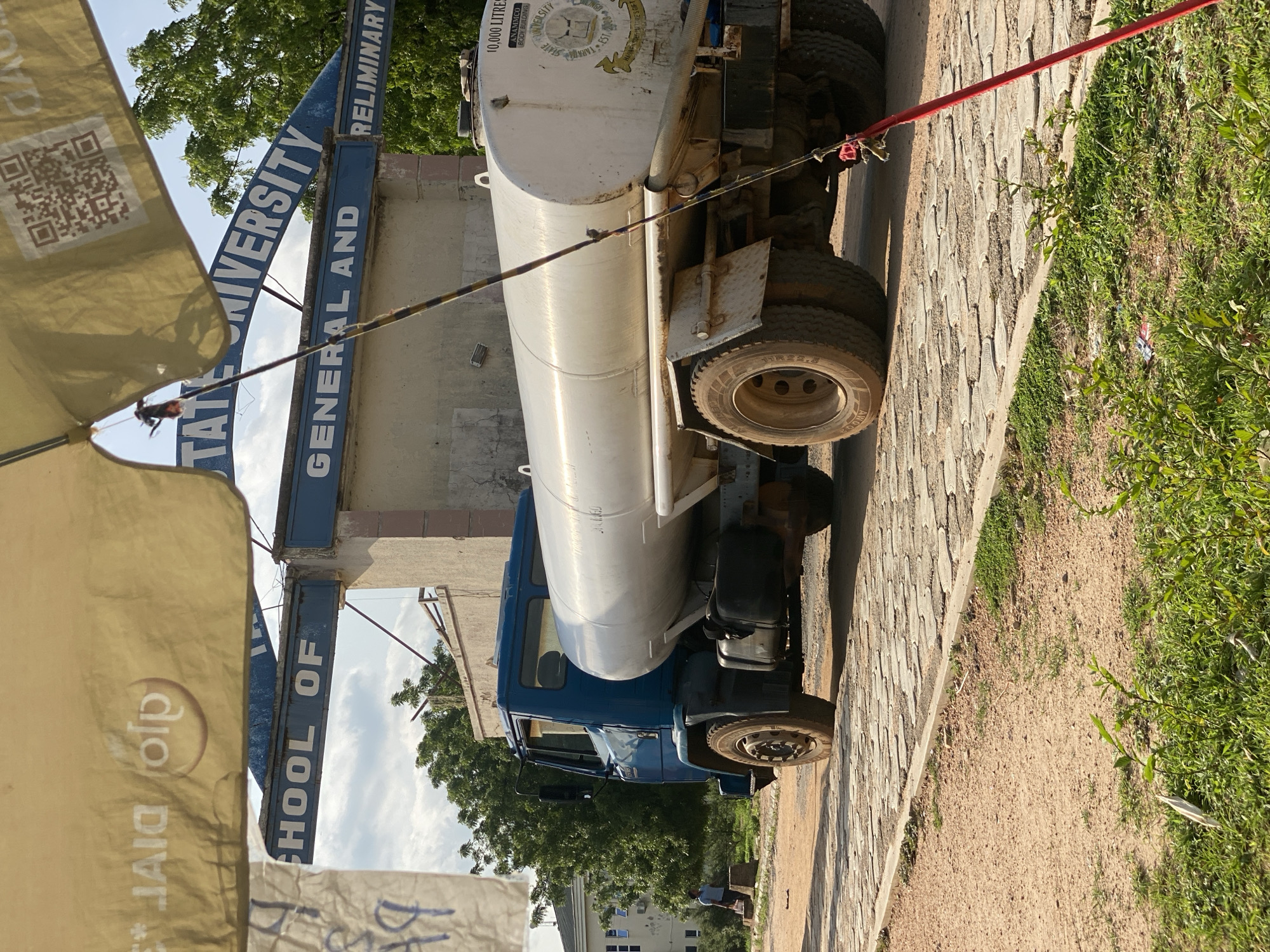 An outdoor, medium shot captures a blue tanker truck parked on a cobblestone path in Jalingo, Nigeria. The truck is positioned diagonally across the frame, with its large, silver tank prominently displayed. The tank has text on its rear, partially visible, including "10,000 LITRES" and a crest with "TARABA STATE UNIVERSITY EST. 2006". The truck is parked in front of a light-colored building with a blue sign that reads "SCHOOL OF GENERAL AND PRELIMINARY". In the background, lush green trees frame the scene under a cloudy sky. A yellow canopy extends from the top of the frame, obscuring the top portion of the building and signage. A single person, dressed in a blue shirt, is visible walking in the distance on the left side of the image. A red pole, likely part of the canopy structure, extends from the right side of the frame downwards. The ground is a mix of dirt and cobblestones, with patches of green grass growing along the edge of the path. The lighting suggests it is daytime, with shadows cast on the ground.