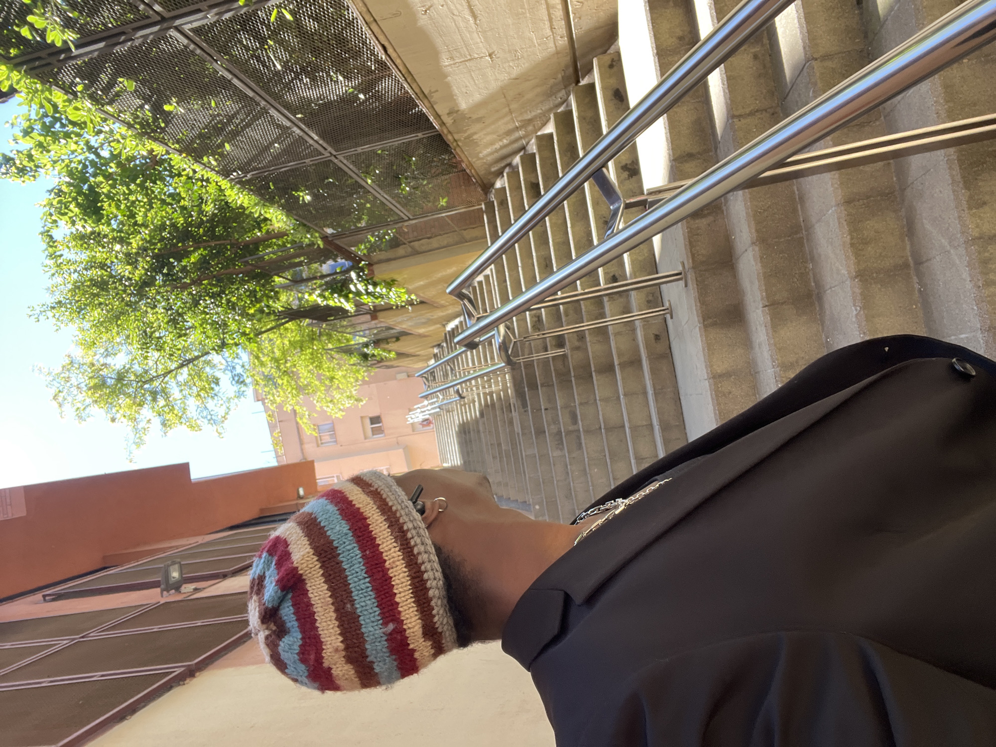 The image shows a person wearing a colorful knitted beanie and a black jacket, standing next to a set of stairs with metal handrails. The stairs lead up to an area covered by a trellis with green foliage. In the background, buildings are visible under a bright sky. The location appears to be an outdoor urban environment, with architectural features typical of Barcelona, Spain, suggesting a possible location within the city. The person is looking upwards toward the foliage and buildings.