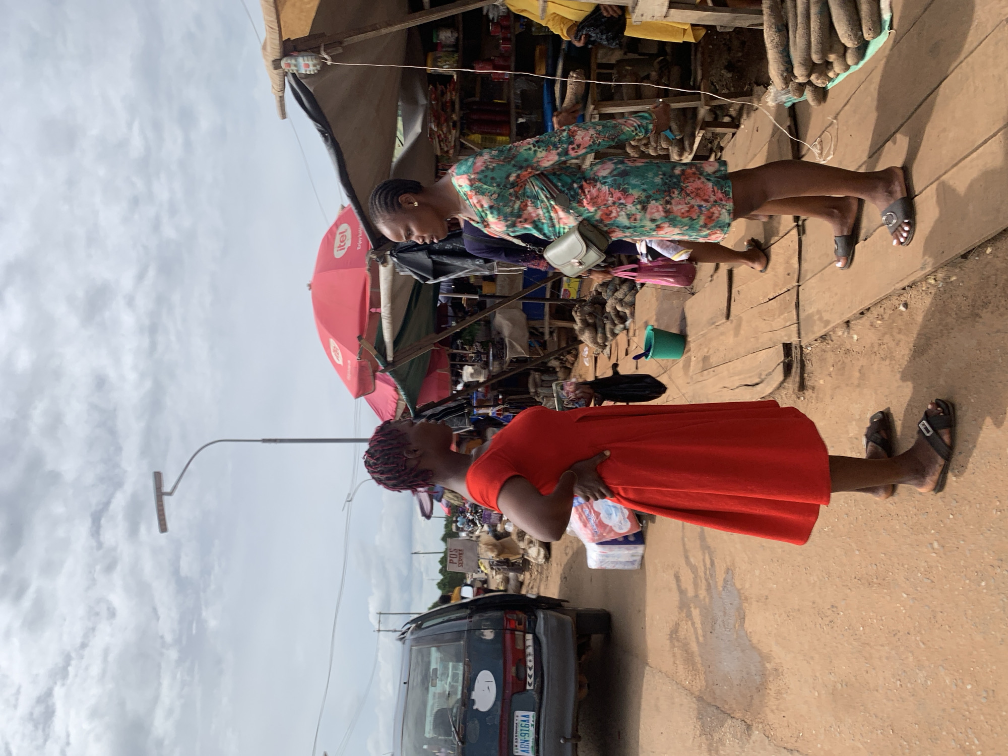 The image shows a street scene in Nsugbe, Nigeria. Two women are prominent in the foreground. One woman is wearing a red dress and sandals. The other woman is wearing a floral dress and sandals and carries a small purse. They appear to be looking up at something out of frame. A dark-colored van is parked nearby. A red umbrella with the word "itel" printed on it is visible. There is market activity visible with various stands and goods displayed. A stack of yams is near one of the stalls. The sky is overcast. A street light pole is visible. There's a sign with the words "POS SERVICE".