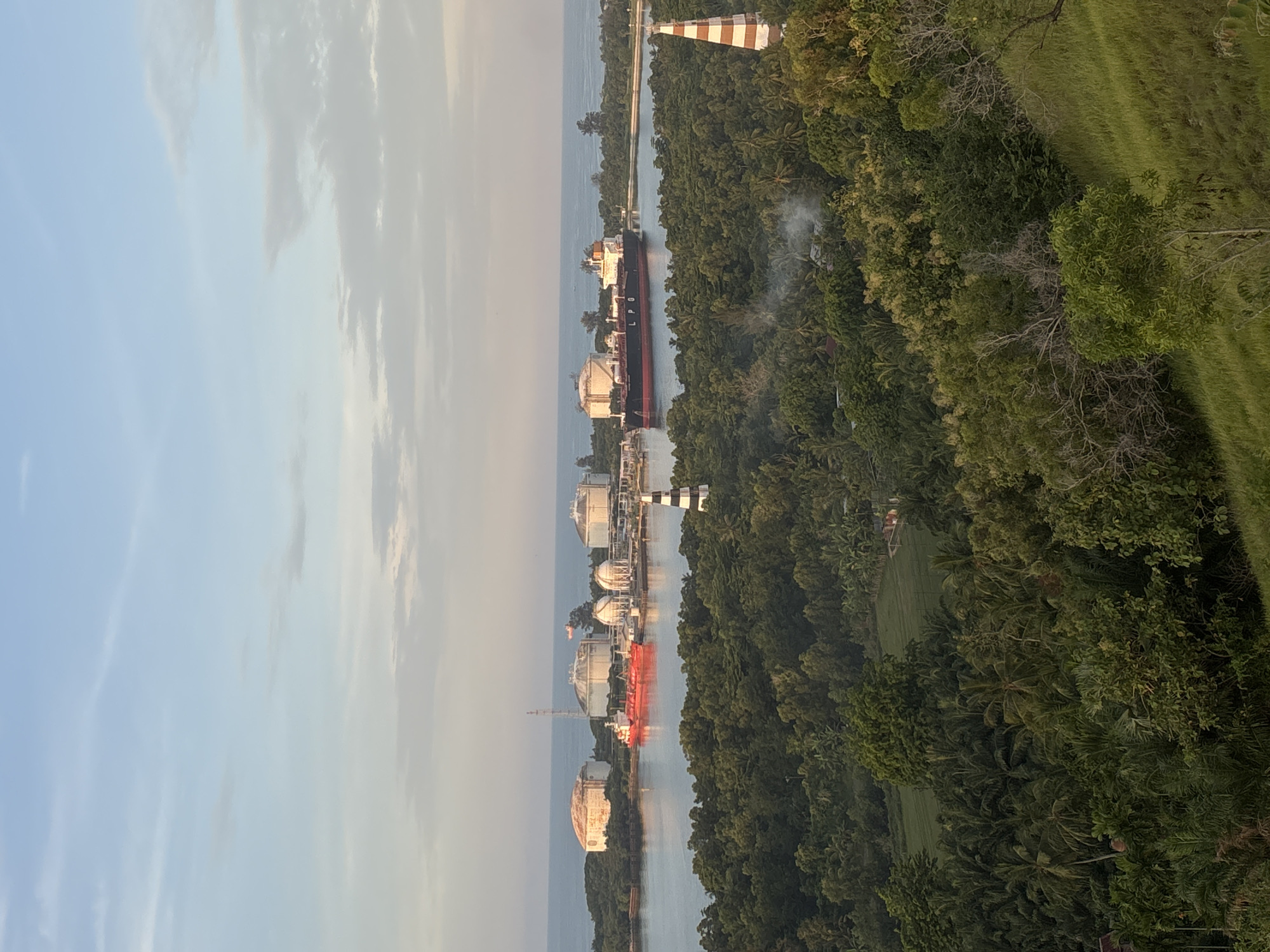 The image shows a coastal landscape in Lhokseumawe, Indonesia. Several large spherical storage tanks are positioned along the water's edge, adjacent to the canal that leads to the sea. Ships and a striped lighthouse are docked near the storage tanks. Lush green vegetation and trees cover the land. The sky is light blue and pale.