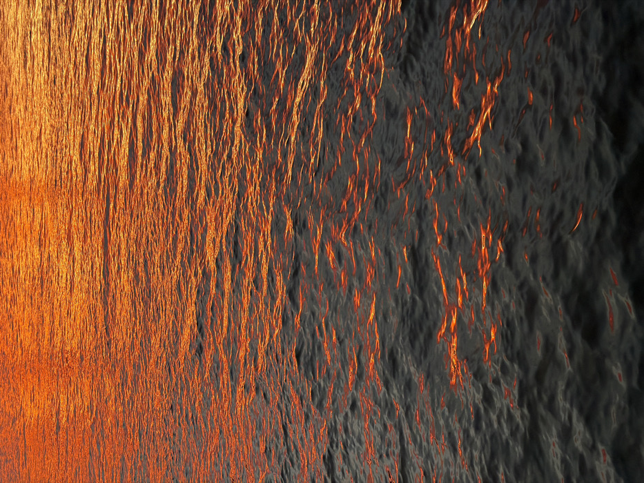 The image displays a detailed view of a body of water, located in the city of New York, United States of America. The water's surface is illuminated by low-angle sunlight, consistent with either sunrise or sunset conditions. The upper two-thirds of the frame feature widespread, intense orange and red reflections, forming numerous elongated, undulating light streaks across the water's rippled texture. The lower third of the frame shows a transition to darker, deep grey-black water, where reflections of orange and red light appear more scattered, fragmented, and less intense. No individuals, objects, or structures are visible within the frame. The scene primarily depicts the dynamic interaction of light reflection with the moving water surface.