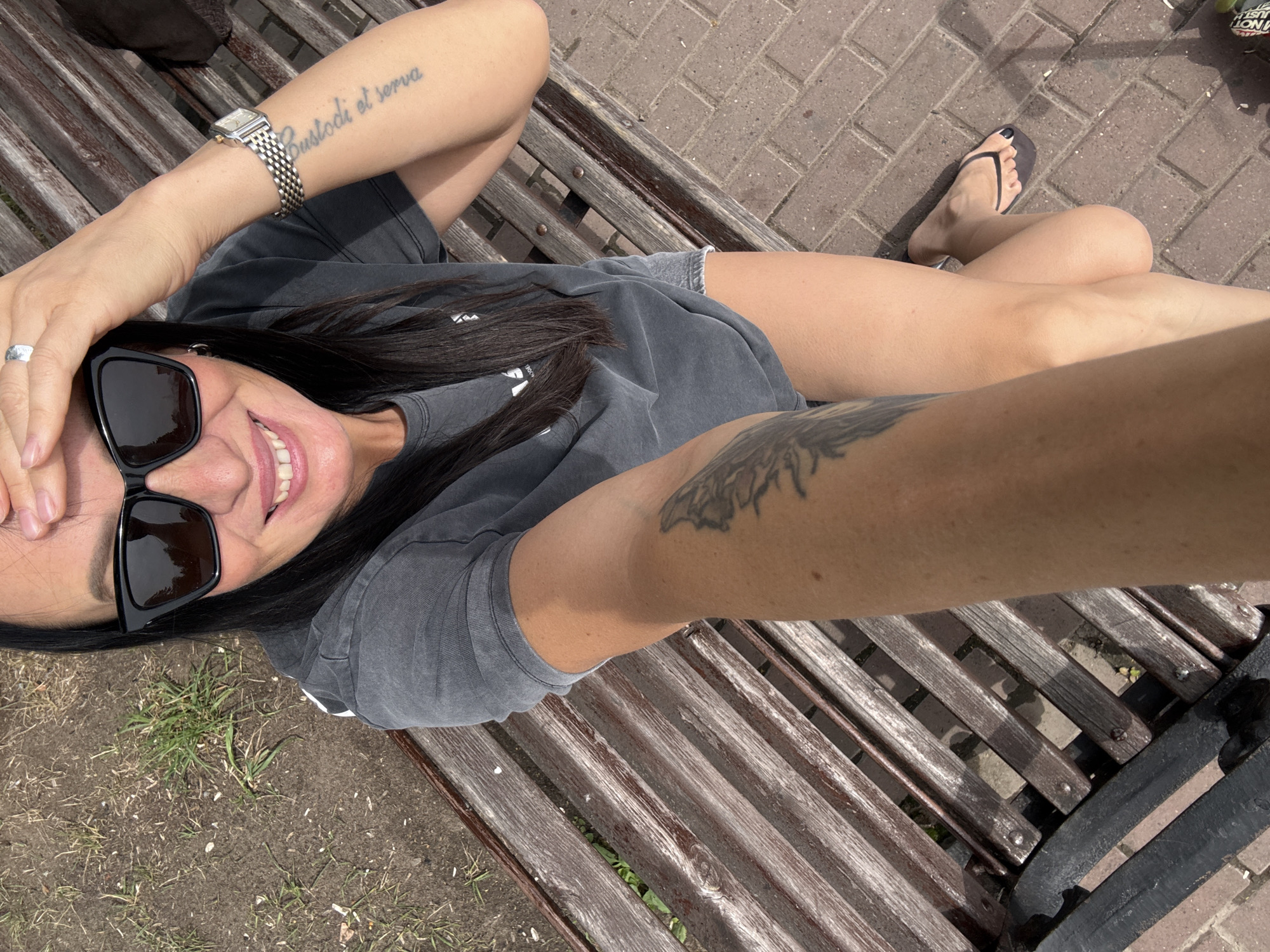 A woman wearing sunglasses and a dark t-shirt is sitting on a wooden bench on a sunny day. She is taking a selfie with her left arm extended. A tattoo is visible on her left arm. A watch is on her right wrist. Her right leg is bent and her right foot is visible, wearing a flip-flop. The bench is made of wooden slats and is placed on a brick walkway. There is some grass and dirt next to the bench. The image appears to have been taken outdoors.