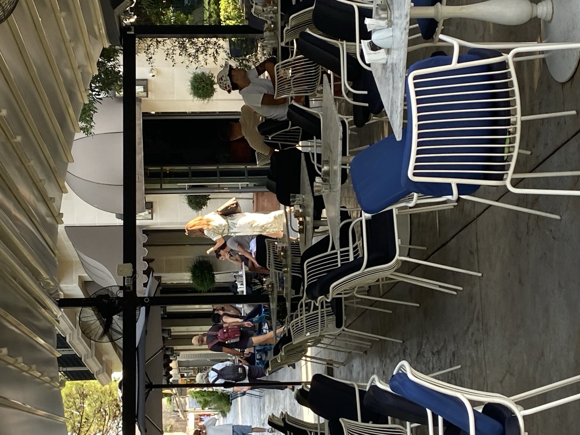 The image depicts an outdoor seating area of a cafe or restaurant in Budva, Montenegro, possibly on the promenade near the Old Town. The scene is characterized by numerous tables and chairs set up on a concrete patio, shaded by a retractable awning. In the background, a building with light-colored stone facade and awnings can be seen, suggesting an urban setting.
Several people are present. In the foreground, white metal chairs with blue cushions are arranged around marble-topped tables. Further back, people are seated at tables, some appearing to be dining or conversing. One woman in a light-colored dress is standing and looking towards the left. A man with a backpack walks away from the camera, while another man wearing a hat sits at a table. The atmosphere appears relaxed and casual, with natural light illuminating the scene. The presence of fans mounted on poles indicates a warm climate. The overall impression is one of a lively outdoor cafe.