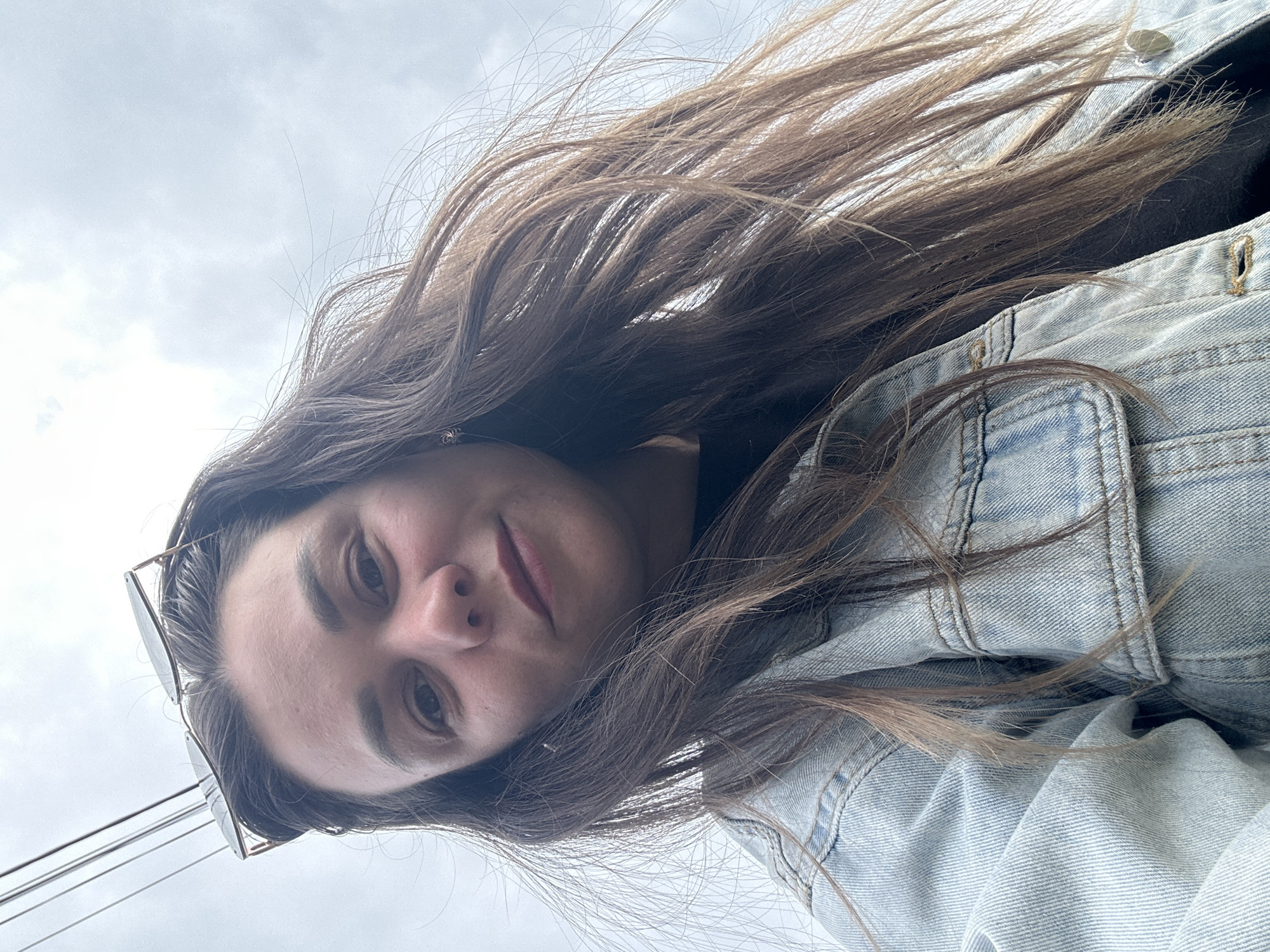 A low-angle, close-up, outdoor, selfie-style portrait shows a woman from the chest up, against a cloudy sky. The woman has long, brown hair that is windswept and falling around her face and shoulders. She is wearing a light-wash denim jacket. A pair of sunglasses is perched on top of her head, resting on her hair. Her right eye is partially obscured by her hair. The background consists of a light gray, overcast sky with faint vertical lines that may be power lines. No specific landmarks in Chaikovsky, Russia are visible.