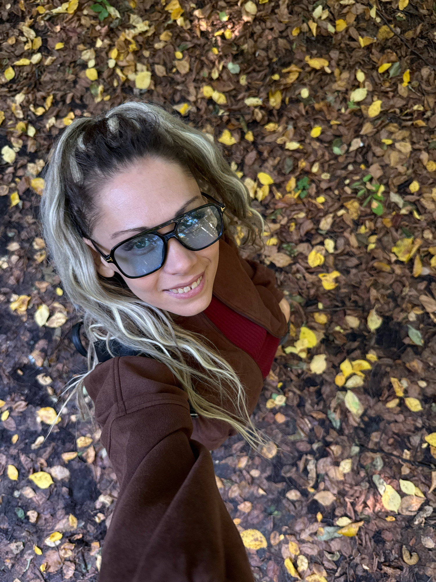 A woman with blonde hair pulled into a high ponytail, wearing a brown jacket over a maroon top and large black-framed sunglasses, is taking a selfie outdoors. She is smiling and looking up towards the camera. The ground around her is covered in fallen autumn leaves, predominantly brown and yellow. The background consists of more leaves and appears to be a wooded or park-like area. The location is the city of Rusanovo, Russia.