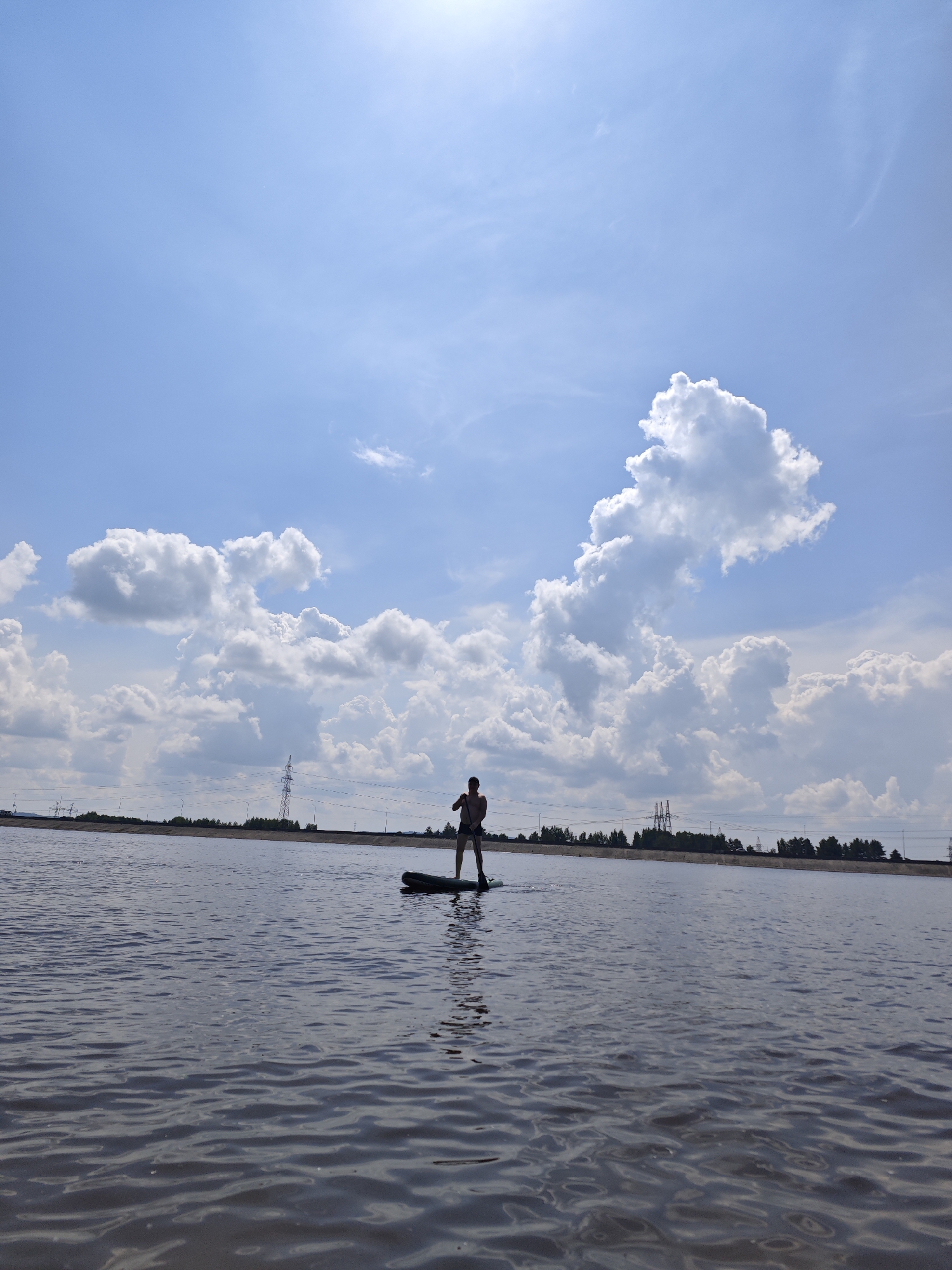 In Noviy, Russia, a person is paddleboarding on a body of water under a bright, cloudy sky. The individual, silhouetted against the sky, stands on a paddleboard and is actively paddling. The background features a flat, treeline along the horizon, with several tall electrical towers visible. The water's surface is rippled, reflecting the cloudy sky. The sun is high, casting a glare on the upper portion of the image.