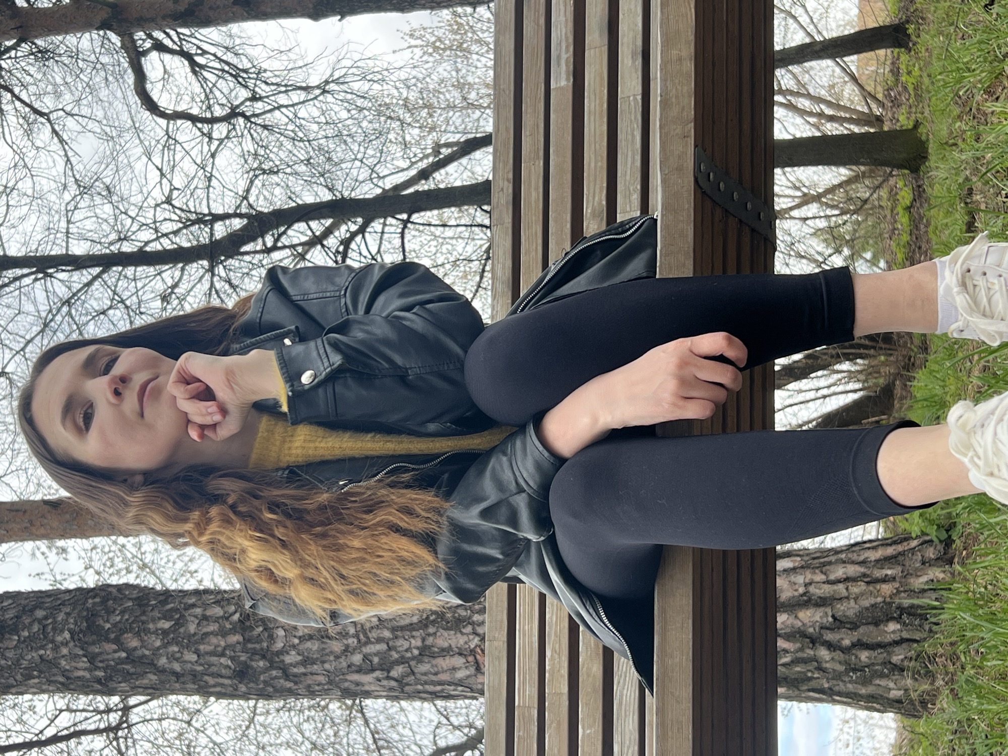 A young woman is seated on a wooden slatted bench in an outdoor park or natural setting in Votkinsk, Russia. The image captures her looking directly at the viewer with a calm, somewhat pensive expression, her left hand resting near her chin.
She has long, wavy brown hair with lighter highlights, which cascades down her back and over her shoulders. She is dressed in a black leather jacket worn open, revealing a mustard yellow knitted sweater underneath. Her attire also includes black leggings, white ankle socks, and white sneakers.
The background is dominated by numerous bare tree branches against an overcast sky, indicating either late winter or early spring. A thick tree trunk is visible to the left of the woman, and green grass can be seen on the ground to the right of the bench. The wooden bench features vertical slats for the backrest and horizontal planks for the seat, with dark metal supports. The lighting is soft and diffused, consistent with an overcast daytime. No text is visible in the image.