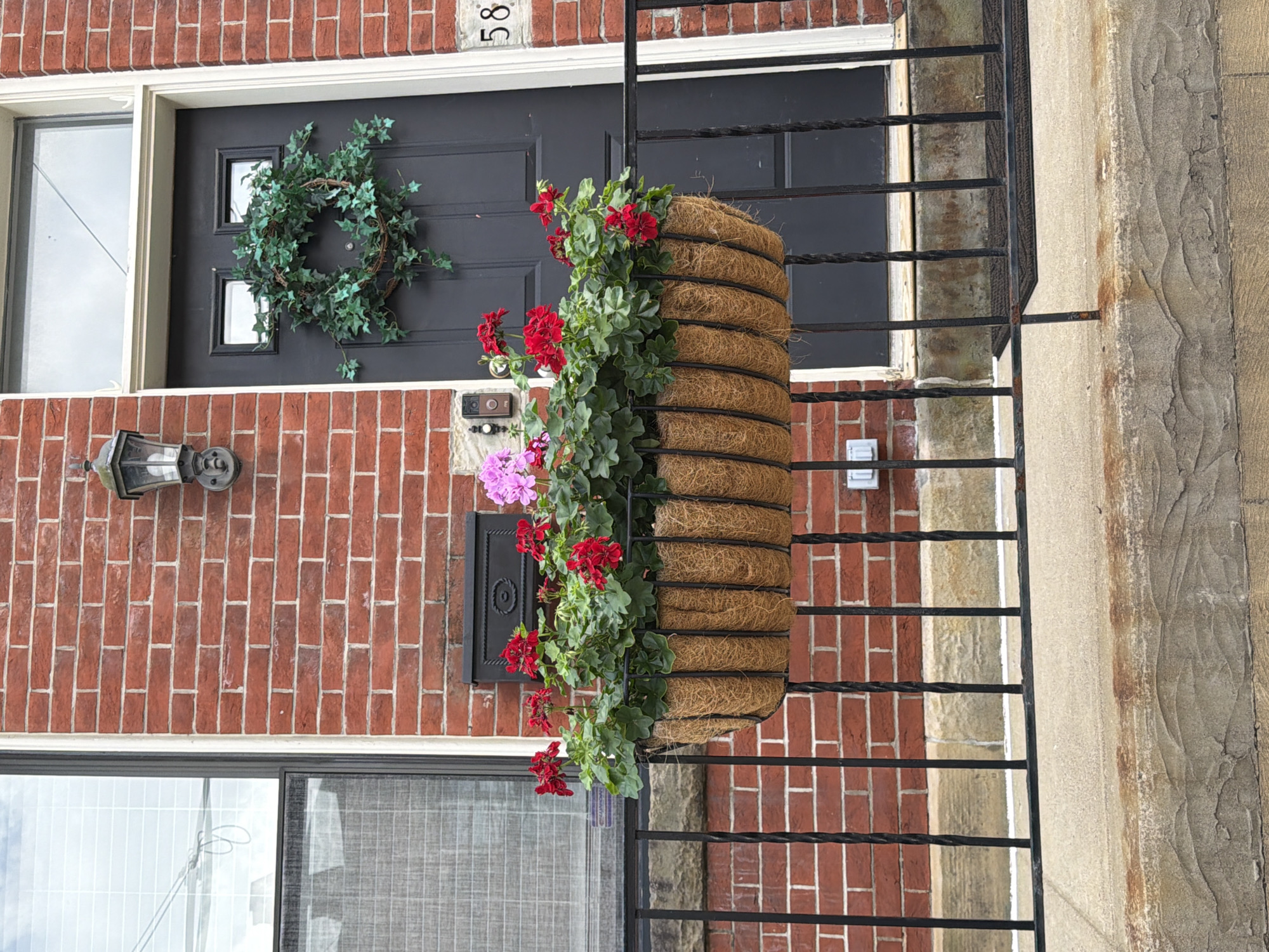 The image depicts the exterior of a brick building in Pittsburgh, United States of America. The building features a dark gray front door adorned with a green ivy wreath. To the left of the door is a window with blinds. A black wall-mounted lantern is positioned above the door. In front of the building, a black metal railing encloses a porch area. A long, oval-shaped planter filled with red and pink flowers is attached to the railing. The planter appears to be made of a woven natural material. To the right of the door, the number "58" is visible. The brickwork of the building is a reddish-orange hue. The ground in front of the porch is a light-colored concrete or stone surface, with a dark doormat visible near the door. The scene is captured in daylight.