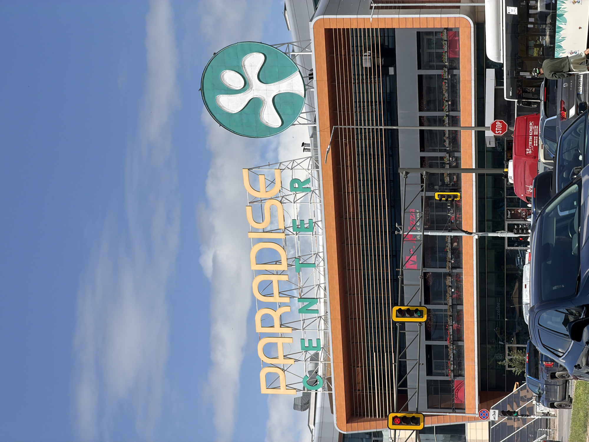 The image shows the exterior of the Paradise Center shopping mall in Sofia, Bulgaria. The mall's signage is prominent, featuring a green circular logo with a white abstract shape and the name "PARADISE CENTER" in yellow and green letters. The building facade is visible, with brown vertical slats and glass windows. Traffic lights are visible along with cars and a red van parked along the street. The sky is blue with white clouds.