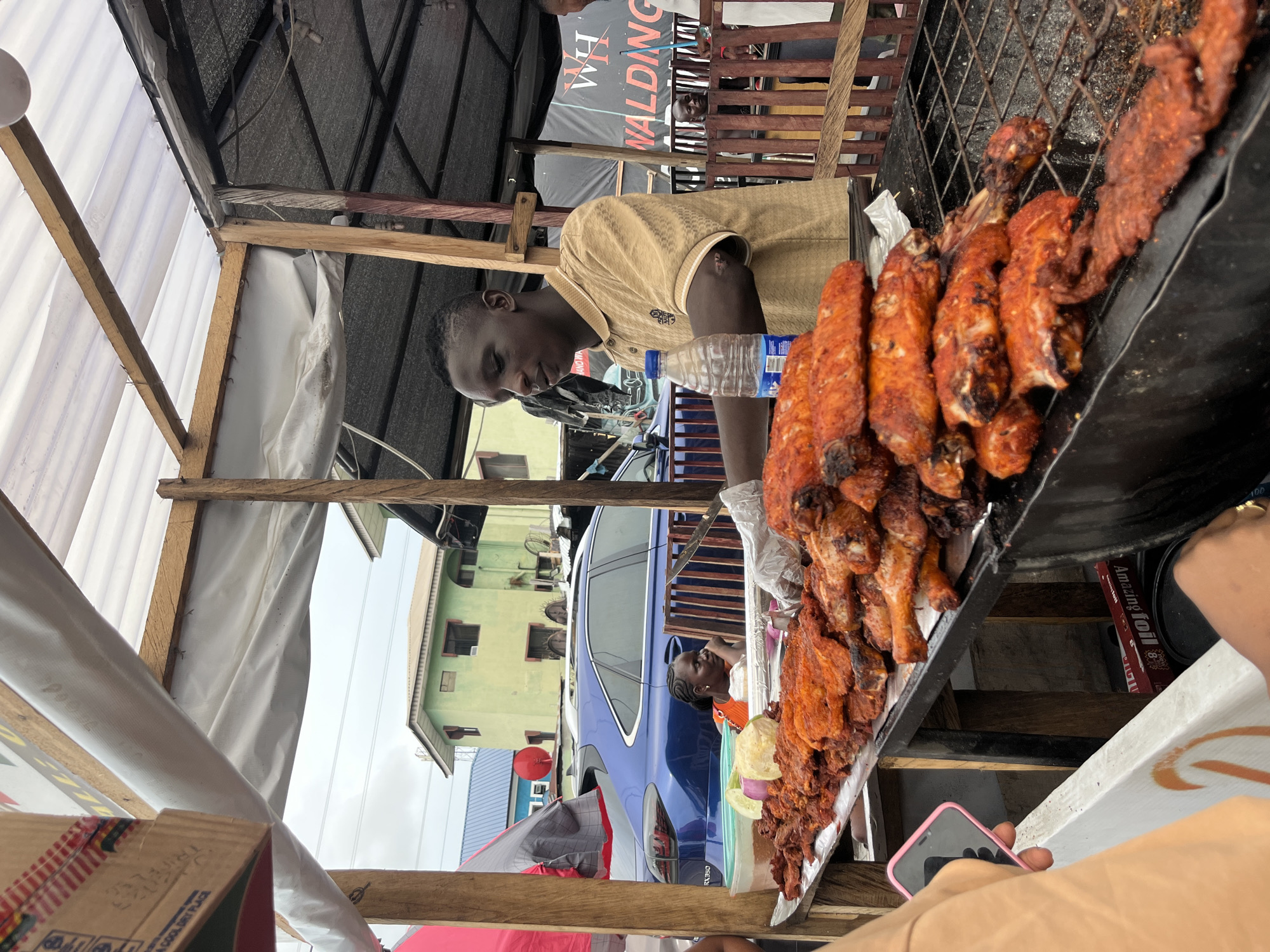 The image captures a street food scene, likely in Ado, Nigeria. A man is standing behind a grill laden with grilled chicken wings and possibly other meat cuts, arranged on a tray for display and sale. He wears a gold-colored shirt and appears to be minding the food and possibly preparing it. A bottle of water rests on the grill counter.
In the background, there is a light blue SUV parked, reflecting the surrounding buildings in its windows. A woman is visible in the background, standing near the car. The environment includes a temporary structure with a white tarp roof supported by wooden beams, indicating a makeshift or temporary food stall setup. A sign with the word "WALDING" is partially visible in the background.