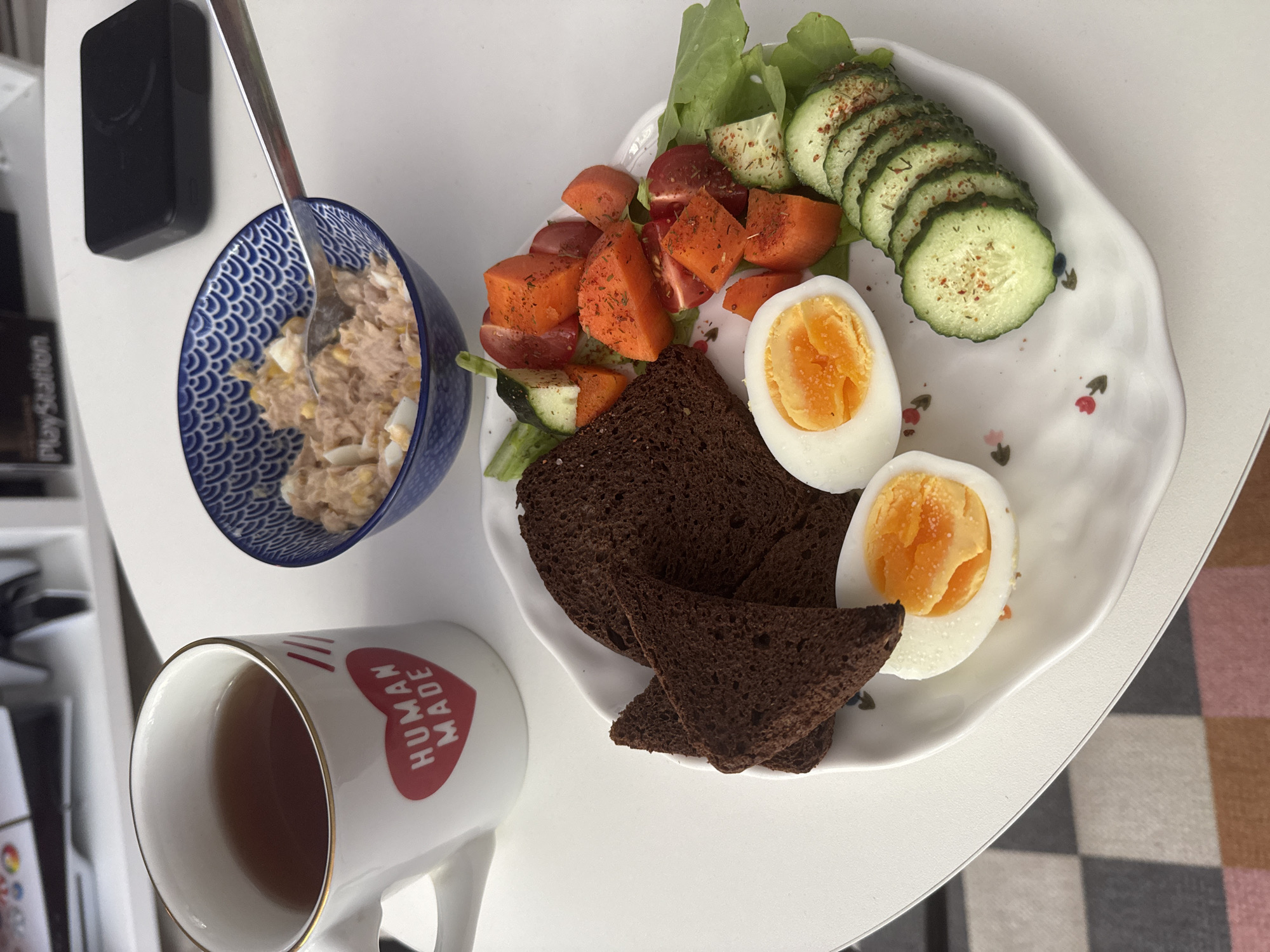 The image shows a close-up, overhead view of a breakfast meal arranged on a white, wavy-edged plate. The meal consists of a salad with lettuce, chopped tomatoes, diced carrots, and sliced cucumbers seasoned with spices. Two halves of a hard-boiled egg, each with a bright orange yolk, are placed on the plate alongside several slices of dark, dense bread. To the left of the plate, a blue and white patterned bowl contains a tuna salad mixed with chopped hard-boiled egg. A silver spoon rests in the bowl. In the foreground, a white ceramic mug with a gold rim is filled with a dark amber liquid, likely tea. The mug features a red heart graphic with the text "HUMAN MADE" printed in white inside. The meal is set on a white surface, possibly a table, with a glimpse of a checkered surface in the background that suggests a home or cafe setting. The lighting is bright, indicating daytime, and there are no visible people. The overall impression is of a healthy and appetizing breakfast.