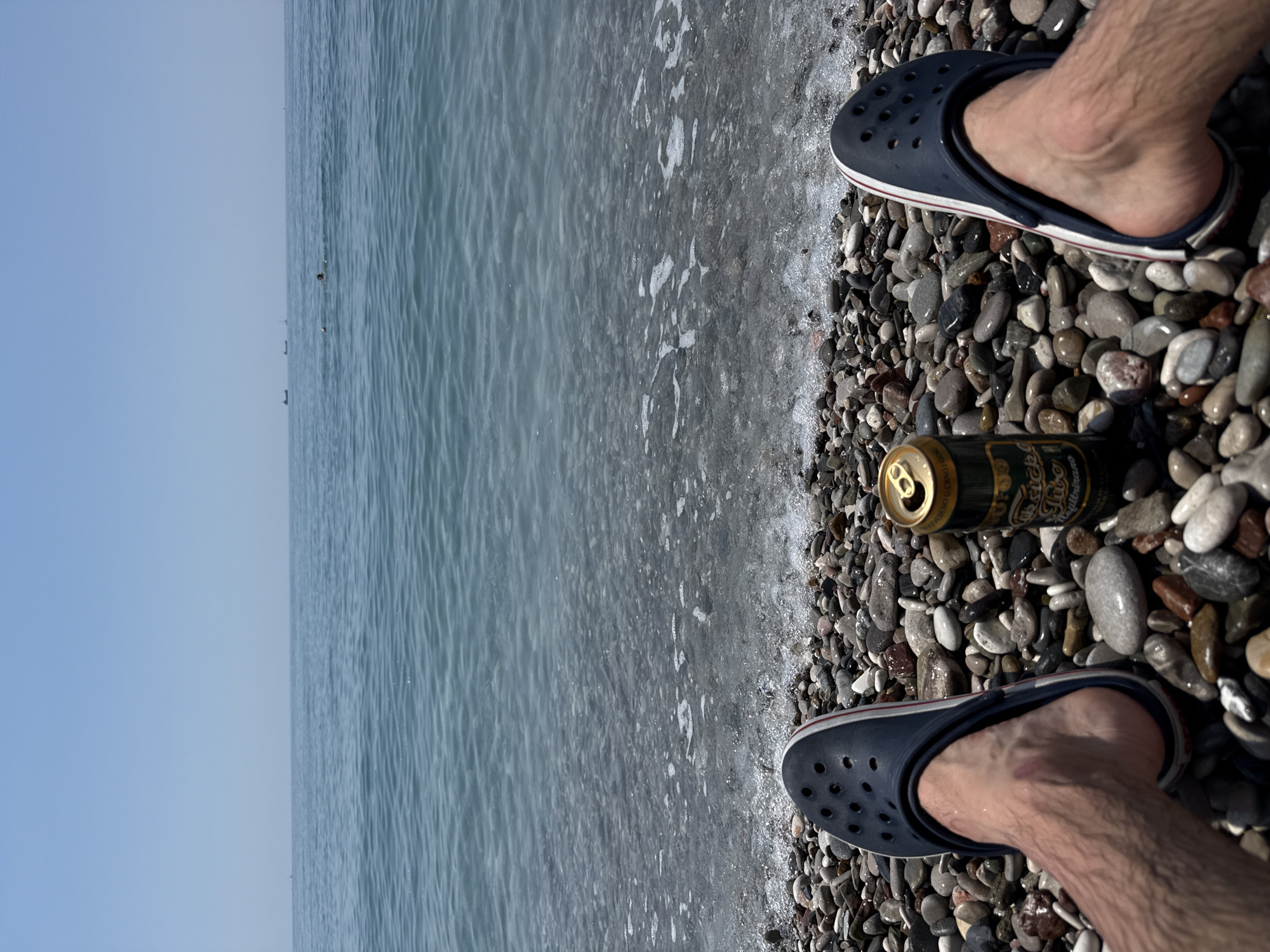 The image depicts a beach scene in Bar, Montenegro, showing a pair of feet in dark blue Crocs standing on a pebble beach. A can of "Niksicko" beer is positioned on the pebbles between the feet. The beach transitions into the sea, and the horizon is visible in the background under a clear sky. Water is lapping around the pebbles near the feet.