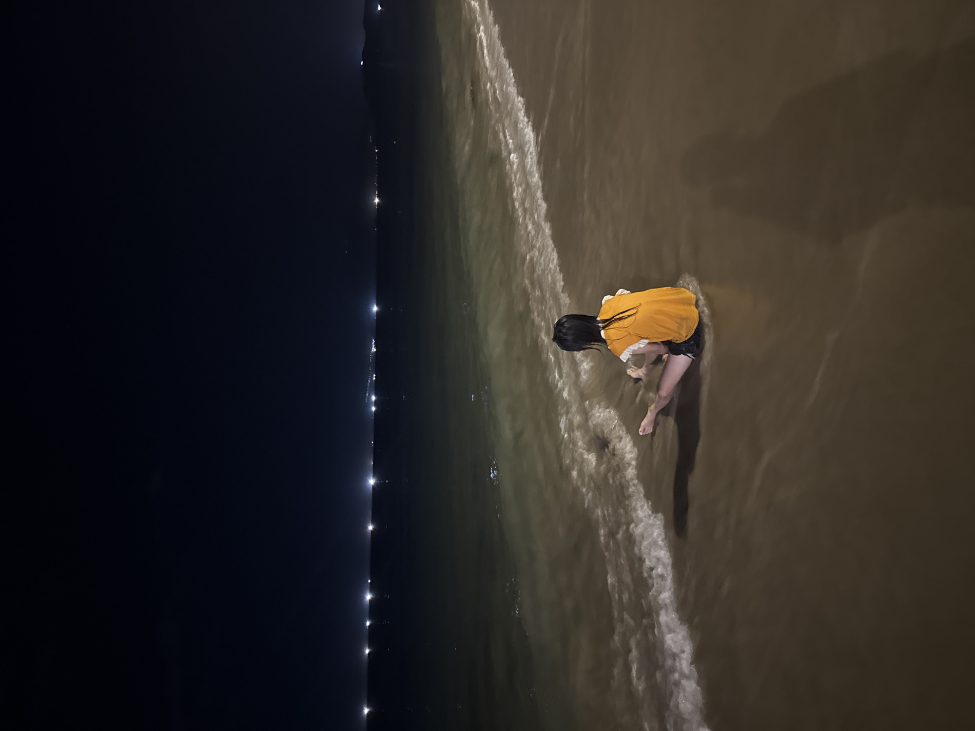 A rear view of a young person with dark hair, wearing an orange t-shirt and dark shorts, sits on a sandy beach at night, facing the ocean. Gentle waves wash over their feet and legs, which are partially submerged in the shallow water. The beach appears wet, and there is a distinct shadow cast on the wet sand to the right of the person. The ocean is dark, with a line of distant lights shimmering on the horizon, suggesting a coastal city. The sky above is a deep, dark blue.
