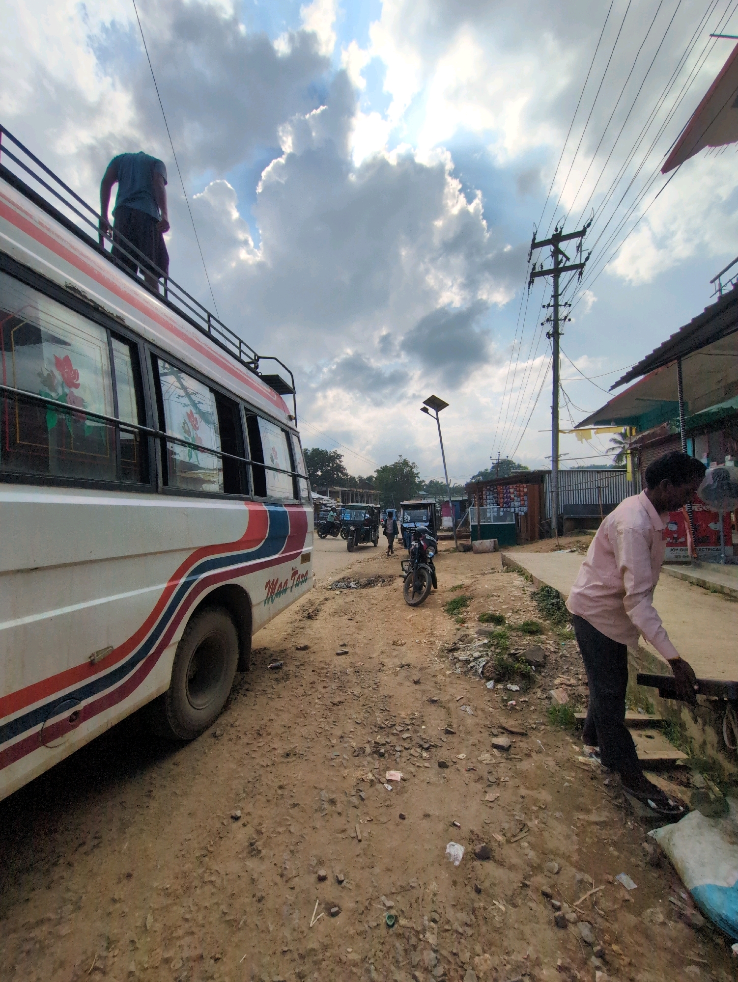The image captures a street scene in Kanchanpur, India, on a cloudy day. In the foreground, a white bus with red and blue striped detailing is parked on a dirt road. A person is standing on the roof of the bus, partially visible. To the right of the bus, a man in a light pink shirt and dark pants is bending over near a sidewalk, appearing to be engaged in an activity by a water pipe. Several motorcycles and auto-rickshaws are present further down the street, indicating vehicular traffic. The background features a row of shops with corrugated metal roofs, a utility pole with numerous wires, and some vegetation. The ground is a mixture of dirt and scattered debris, suggesting an unpaved or developing area. The overall atmosphere is that of a bustling, informal marketplace or transit hub.