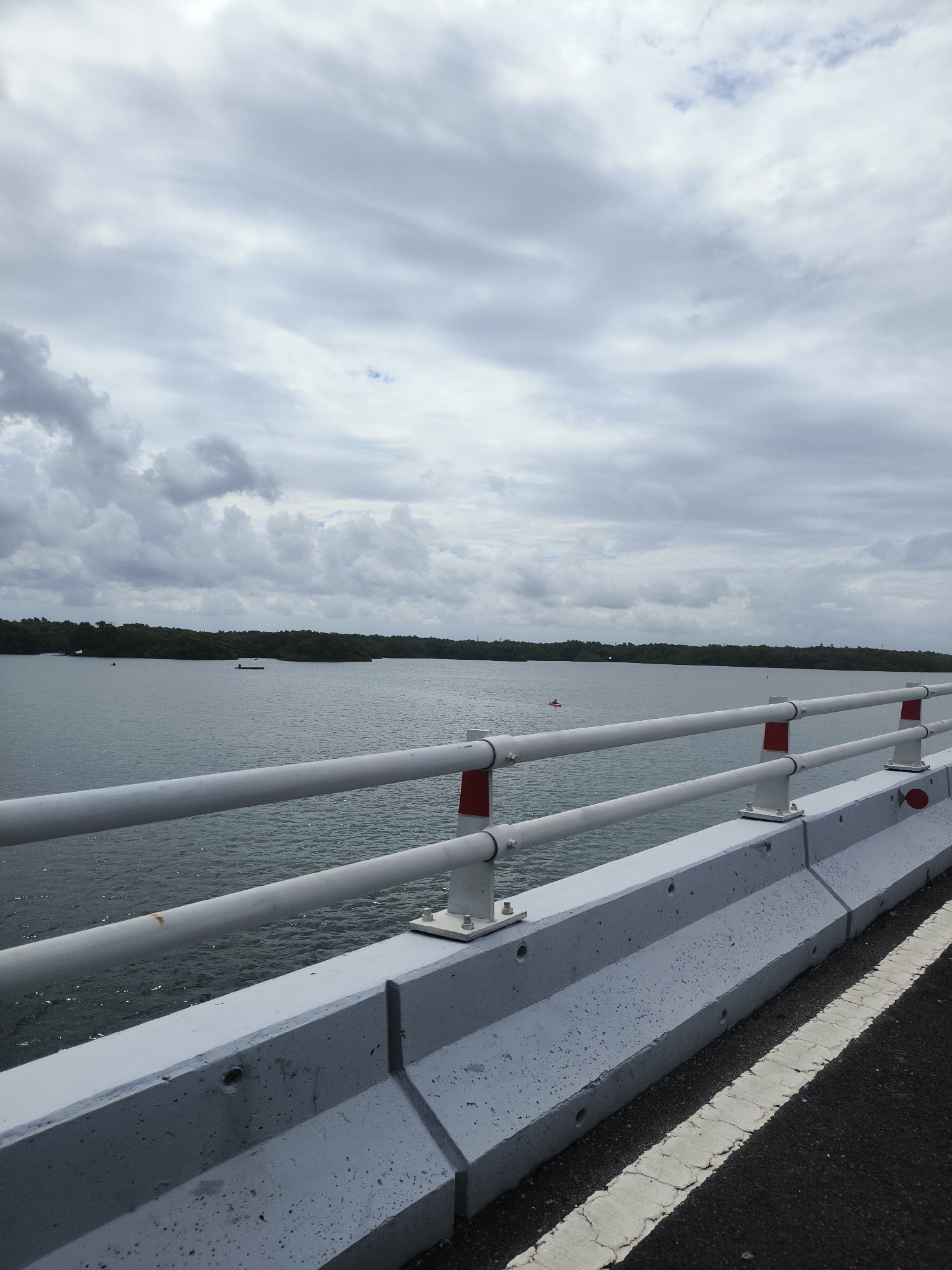 The image depicts a view from a bridge overlooking a body of water in South Denpasar, Indonesia. In the foreground, a concrete barrier lines the side of the road, with a white railing attached to it. The railing consists of two horizontal pipes, supported by posts. The water is calm, with a tree-lined shore visible in the distance. Two small boats are present on the water, one closer to the shore and the other further out. The sky is overcast with a significant amount of cloud cover. The scene appears to be a quiet, possibly rural, area within or near the city.