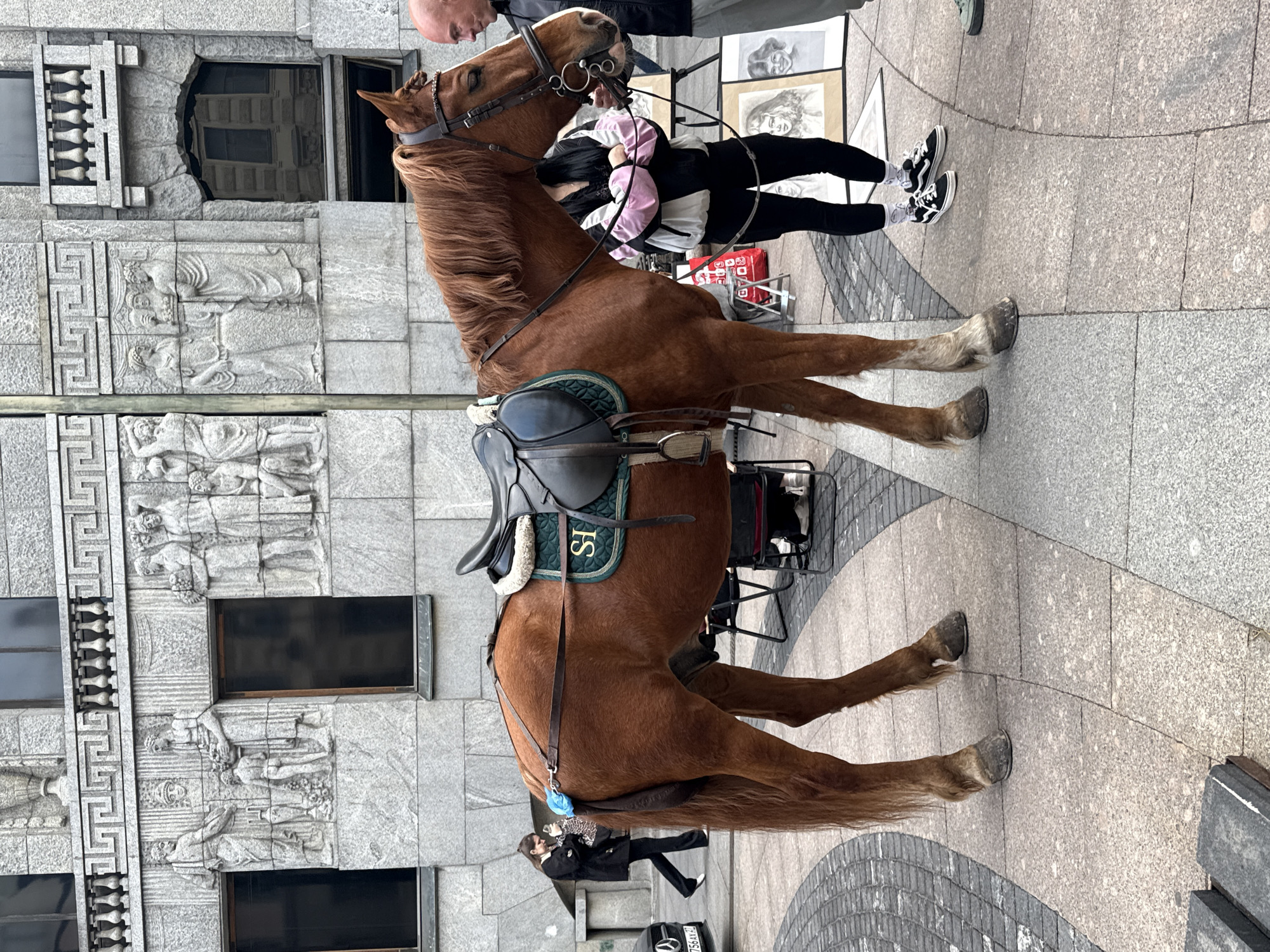 A brown horse stands on a paved street next to a building with intricate stone carvings. The horse is wearing a saddle with green padding and the letters "SI" are visible on it. A person in a black jacket and white shirt is partially visible near the horse's head. In the background, another person, dressed in black pants and a light-colored jacket, is walking away from the camera, with black and white sneakers visible. Several framed artworks are displayed on the ground behind the horse. The scene appears to be in an urban setting, possibly a public square or pedestrian area. The weather seems overcast, as there are no strong shadows. The architecture of the building suggests a historical or European style.