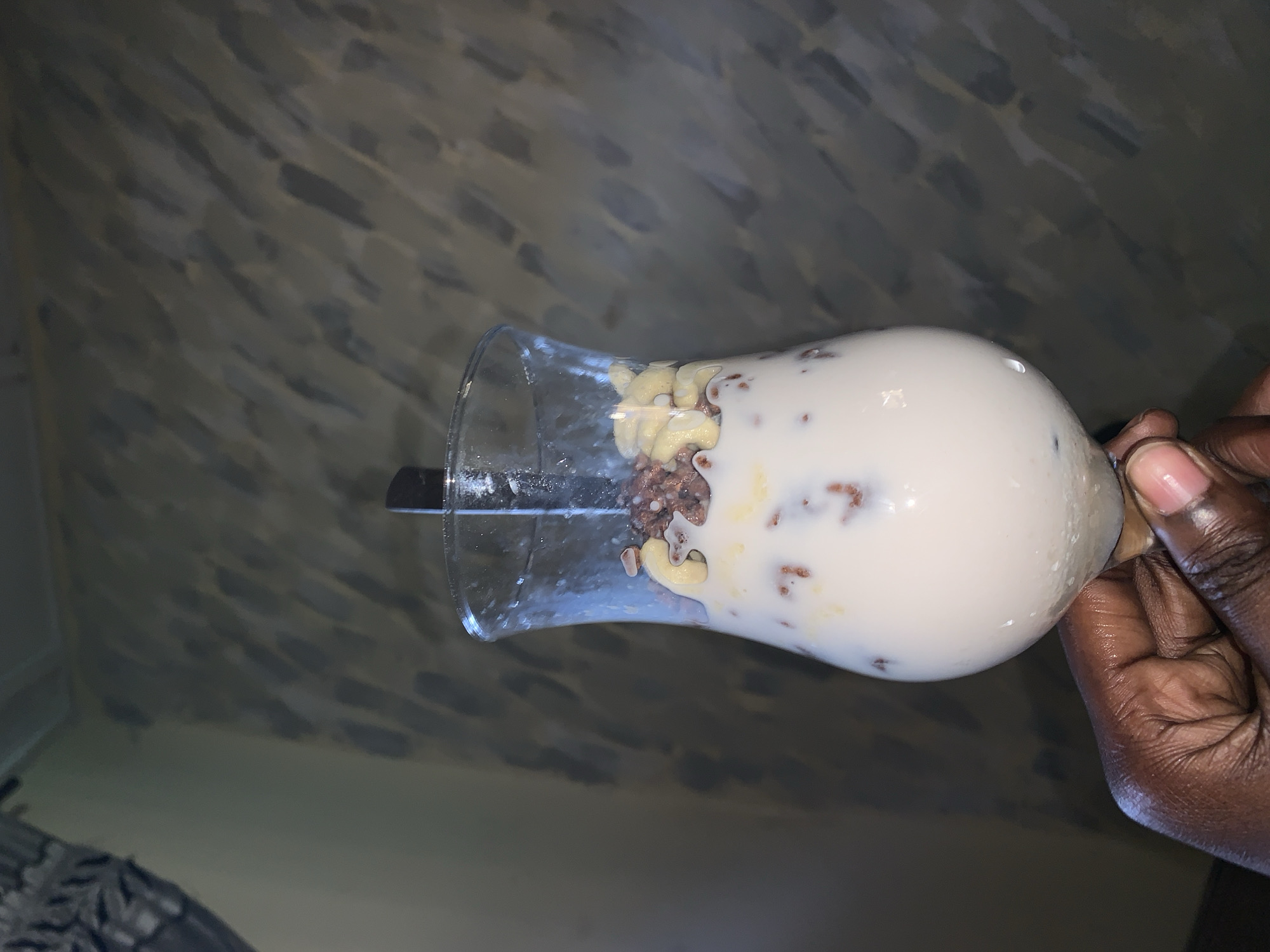 A person holds a glass containing a white liquid, dark grains, and cooked pasta. The image is taken indoors against a wall with a textured pattern. The location, Rafin Par, Nigeria, is not evident in the image.