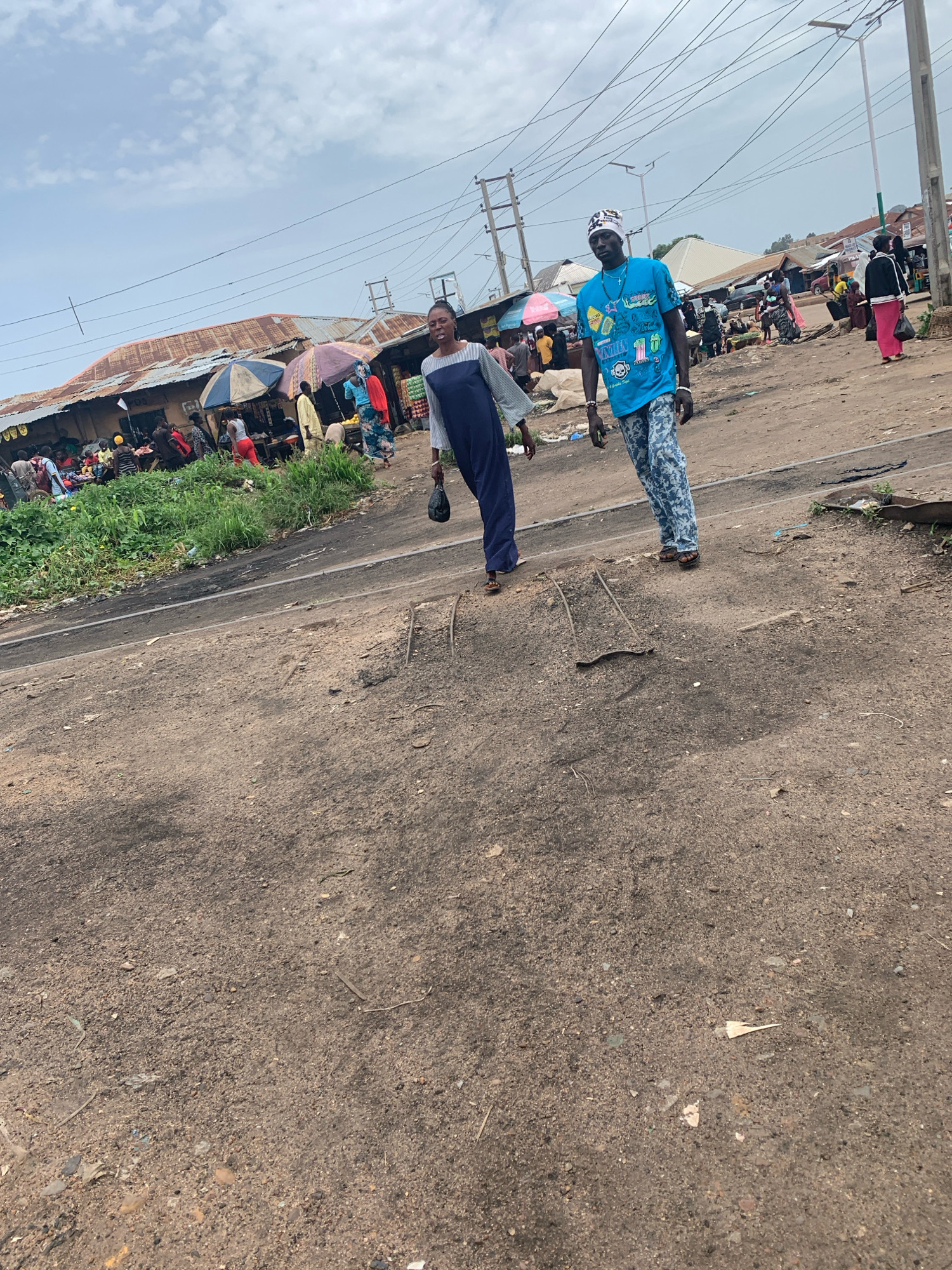 In Bukuru, Nigeria, a dirt road runs through a bustling marketplace. Two individuals walk towards the viewer on the road. On the left, a person in a long, dark blue dress and a lighter blue long-sleeved shirt walks with their head down and a black bag in their right hand. On the right, a person wearing a blue t-shirt with various graphic designs and light blue, patterned pants walks with a slight smile. They are wearing a white and black checkered beanie and brown sandals. In the background, several market stalls with corrugated metal roofs are visible, some adorned with colorful umbrellas. People are seen browsing and interacting within the market. Green vegetation lines the edge of the road, contrasting with the dusty ground. Utility poles and overhead wires are present, indicating the presence of electricity infrastructure. The sky is overcast with scattered clouds.