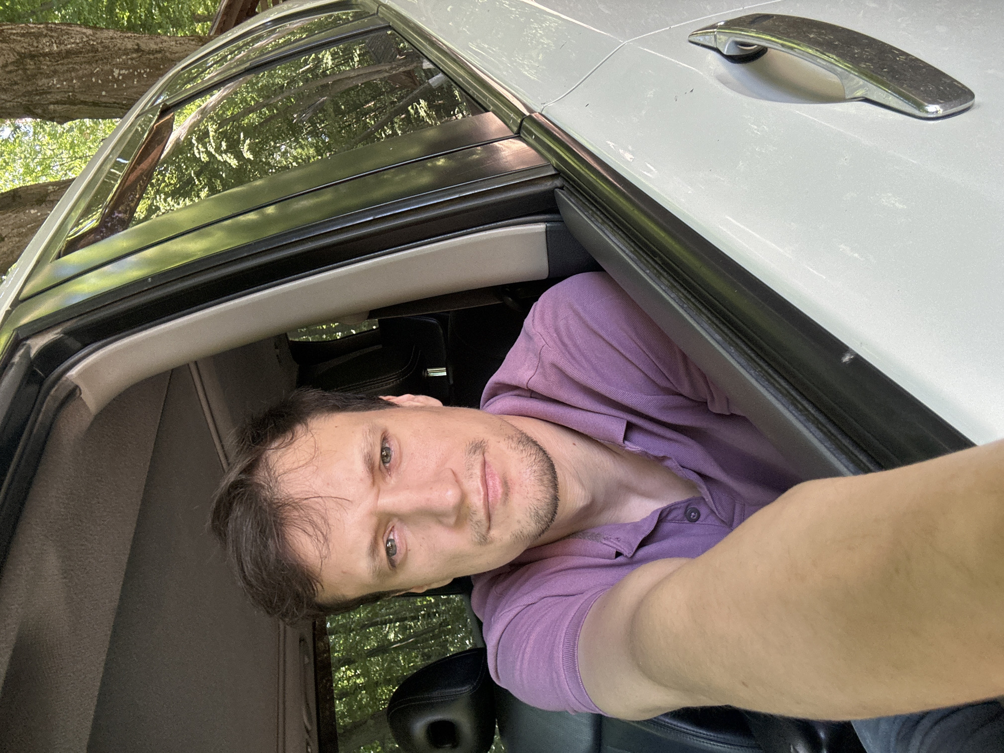 A man with short brown hair and a light beard is taking a selfie from inside a white car. He is wearing a purple polo shirt. The man is positioned in the driver's seat, with his left arm resting on the open car door frame. The interior of the car, including the black leather headrest of the passenger seat, is visible. The background outside the car shows trees and foliage, suggesting a natural setting. The image was taken in Shatava, Ukraine.
