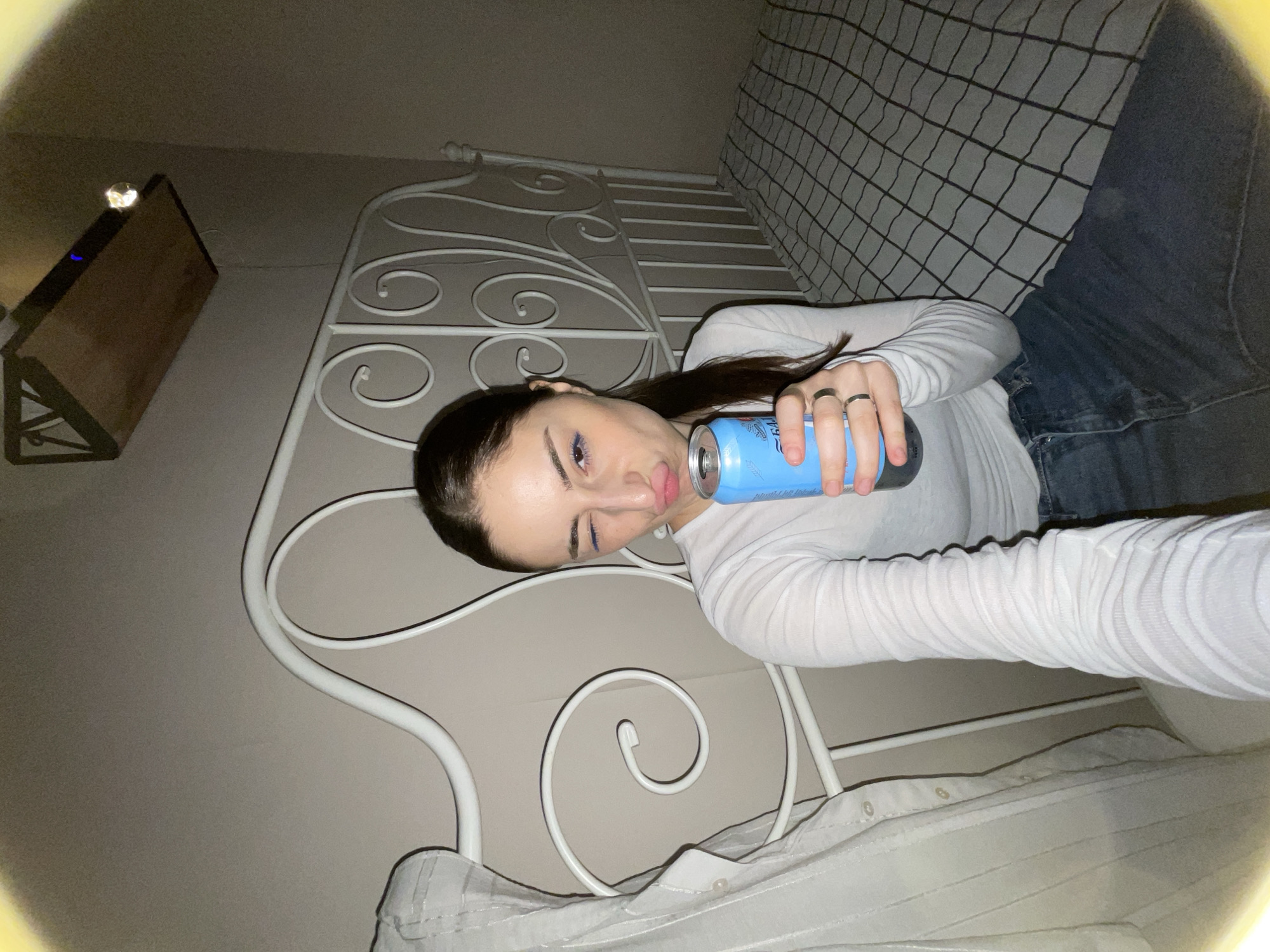 A young woman with dark hair and blue eyeliner is lying on her side on a bed. She is wearing a white long-sleeved shirt and jeans. She is holding a light blue can up to her lips and winking at the camera. The bed has a white metal frame and a gray and white checkered blanket. In the background, there is a light-colored wall and a shelf with a decorative object on it. There is no indication of the city of Moscow, Russia.