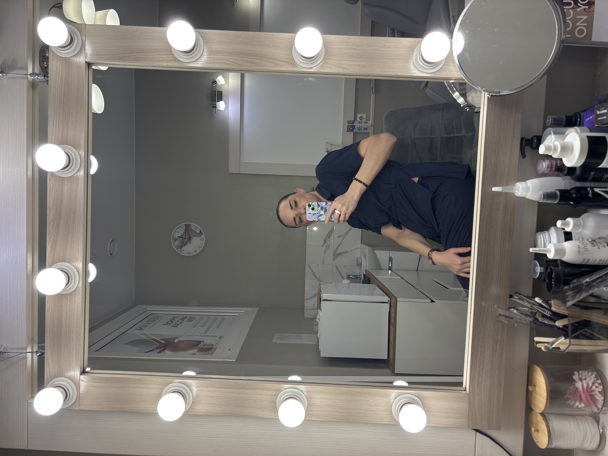 A person wearing a dark blue garment is seen in a vanity mirror. The vanity has lights around its perimeter and a magnifying mirror attached to the right side. Bottles and makeup brushes are visible next to the mirror. In the reflection is a room with white cabinets and tile, a clock on the wall, and a framed picture. It is not possible to determine whether this image was taken in the city of Vsevolozhsk, Russia.