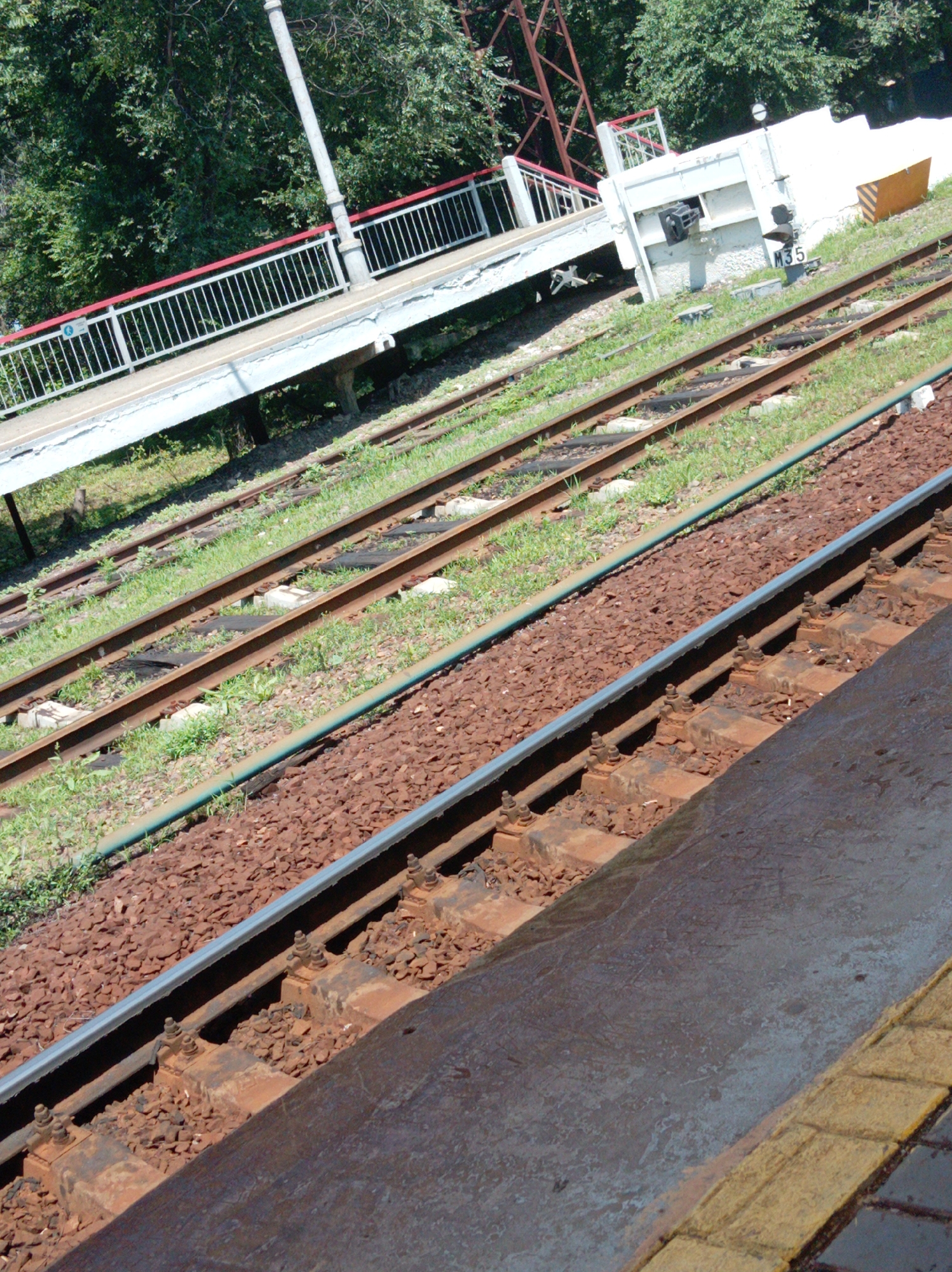 The image shows a section of a railway track with a pedestrian overpass in the background. The railway tracks are made of steel rails laid on concrete sleepers, with crushed stone ballast. The pedestrian overpass is a concrete structure with a metal railing and a ramp leading up to it. In the background, there are trees and foliage. The location is identified as Trudovoye, Russia, a rural settlement. The scene appears to be a sunny day, with the sun casting shadows on the tracks and overpass. There are no people or animals visible in the image. The overall impression is one of a quiet and functional railway infrastructure in a natural setting.