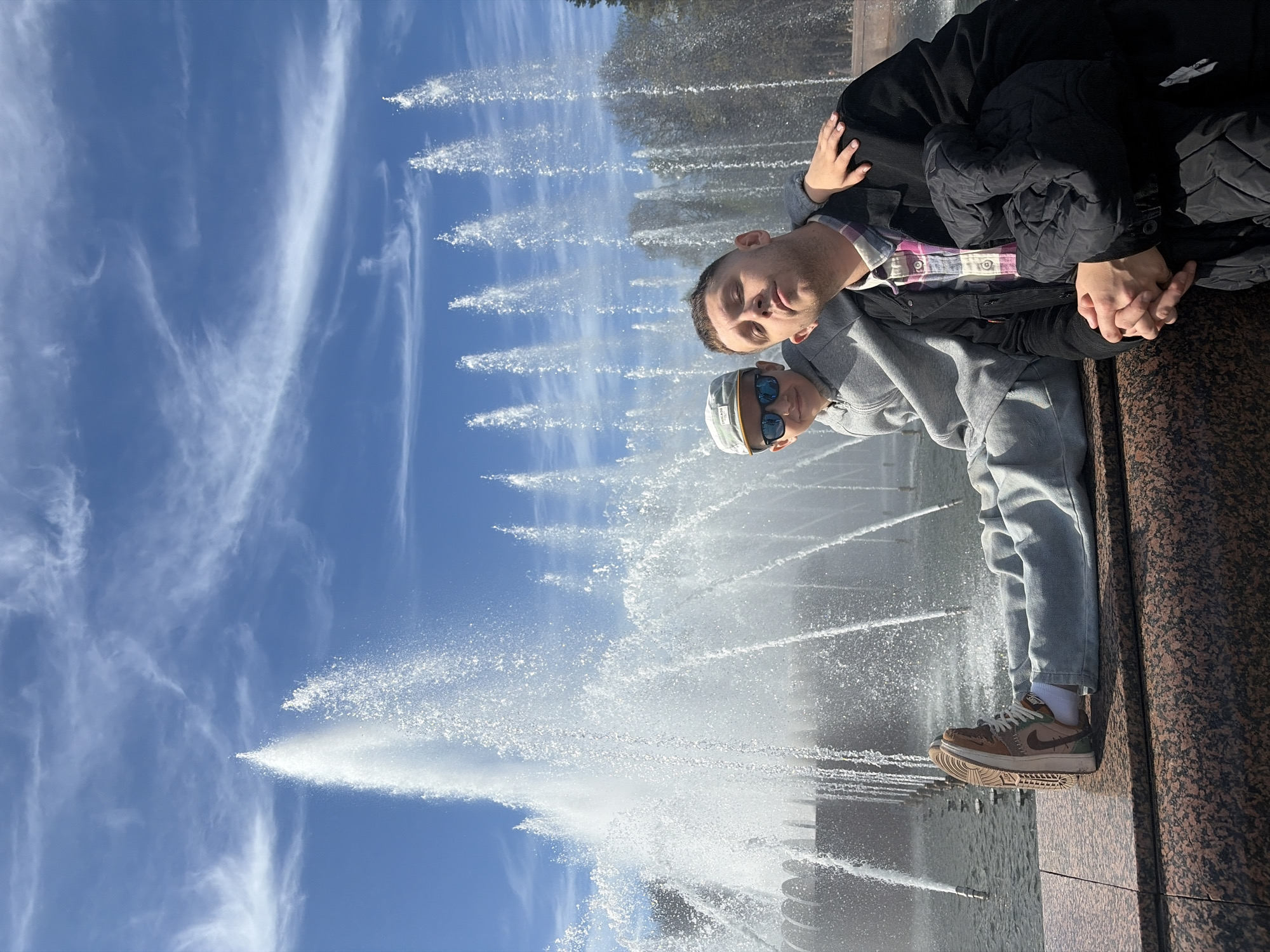 Two men are positioned against a textured brown wall with their legs perpendicular to the ground. One man wears a grey hooded sweatshirt, blue jeans, a white sock, and brown and green tennis shoes. The other man is wearing a light purple and black plaid shirt underneath a black jacket. They are standing in front of a fountain with numerous jets of water spraying upwards. The background consists of a partly cloudy blue sky and an outline of trees. The scene is likely set in Saint Petersburg, Russia, based on the common architecture and fountain designs of the city.