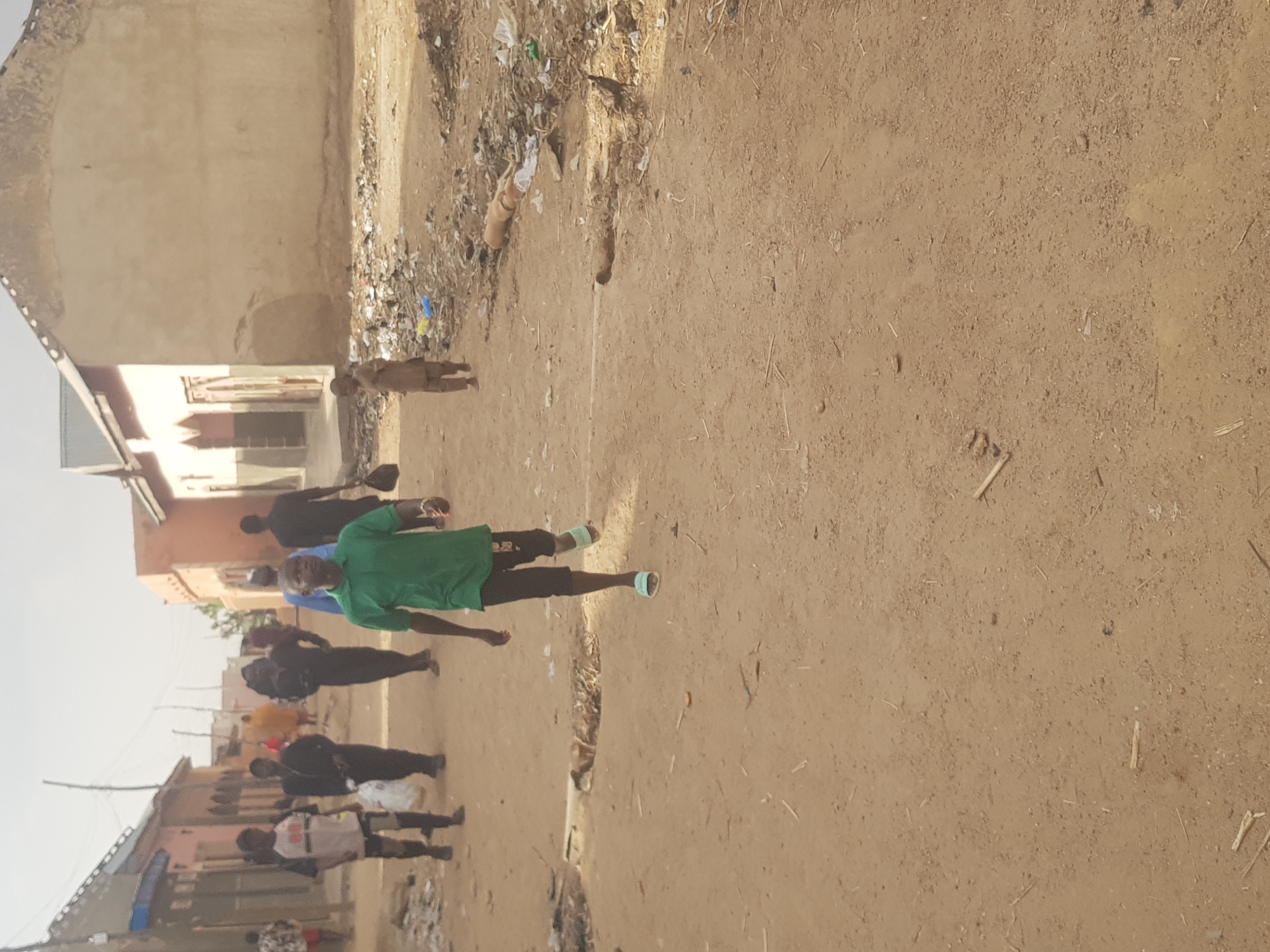In a residential area of Dutsinma B, Nigeria, a group of people are walking along a dirt road. One person in the foreground is wearing a green shirt and dark shorts. Several other people are walking behind them, dressed in casual clothing. The road is unpaved, and there is some trash and debris along the sides. The buildings in the background appear to be simple, one-story structures. The sky is overcast, suggesting it might be early morning or late afternoon.