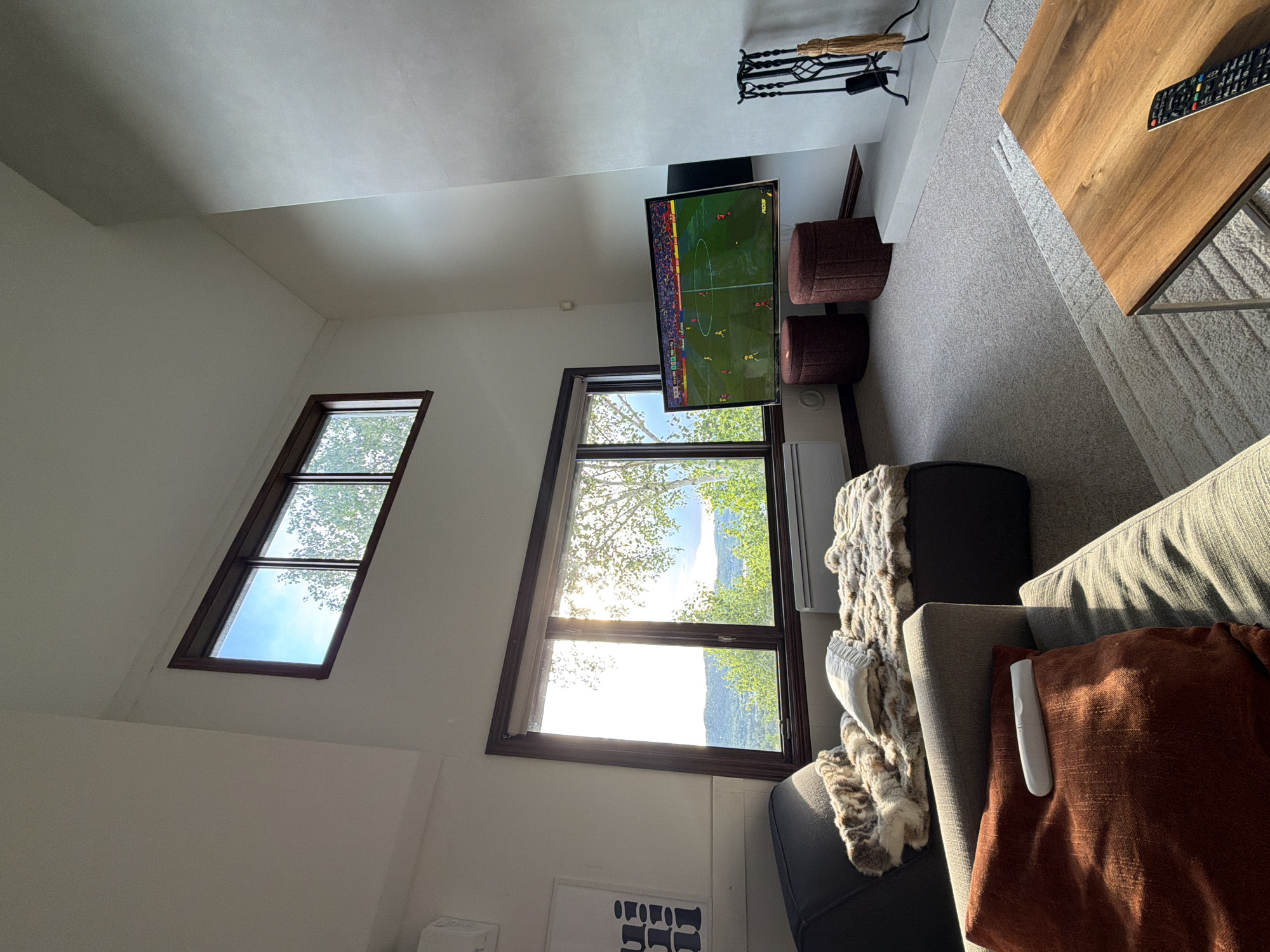 This is a bright, interior shot of a modern living room in a residence in Mont-Tremblant, Canada. The room features a high, vaulted ceiling and large windows that offer a view of lush green trees and rolling hills under a clear blue sky. Sunlight streams in, illuminating the space.
On the left, a dark grey sofa is draped with a plush, patterned throw blanket and a white decorative pillow. In front of it is a dark grey ottoman covered in a similar throw. To the right of the ottoman is a large, wall-mounted flat-screen television displaying a soccer match. Below the TV are two round, burgundy ottomans.
The floor is carpeted in a light grey, textured material. A white fireplace hearth with a black fire iron set is visible to the right. In the foreground, a wooden coffee table with a metal frame holds a black remote control. Another sofa, partially visible in the lower left, is covered with an orange decorative pillow that has a white remote control resting on it. The overall impression is one of a comfortable and contemporary living space with a connection to the natural surroundings.