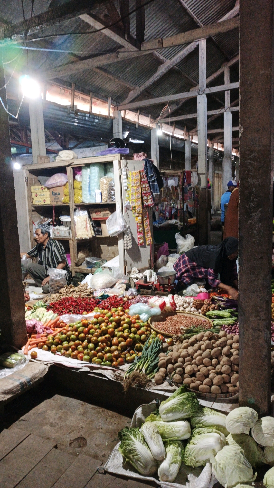 The image depicts a bustling market scene in Cibenda, Indonesia, likely early in the morning or evening due to the artificial lighting. A covered market structure with a corrugated metal roof and wooden beams is visible. Various fresh produce is displayed on the ground and on elevated surfaces.
In the foreground, a variety of vegetables are artfully arranged, including piles of tomatoes, red chilies, carrots, garlic, onions, potatoes, cucumbers, and leafy greens like cabbage and bok choy. There are also baskets and sacks containing nuts and other dry goods.
Two individuals are interacting within the market. On the left, a man wearing a striped shirt and a patterned cap is crouched down, appearing to be engaged in weighing or arranging produce. On the right, a woman wearing a black headscarf and a plaid skirt is also crouched, seemingly tending to the vegetables. Another person in a blue shirt is visible in the background, suggesting other vendors or shoppers.
Shelving units stocked with packaged goods are present in the background, indicating a more permanent stall within the market. The overall atmosphere is one of commerce and local life, with an abundance of fresh food items.