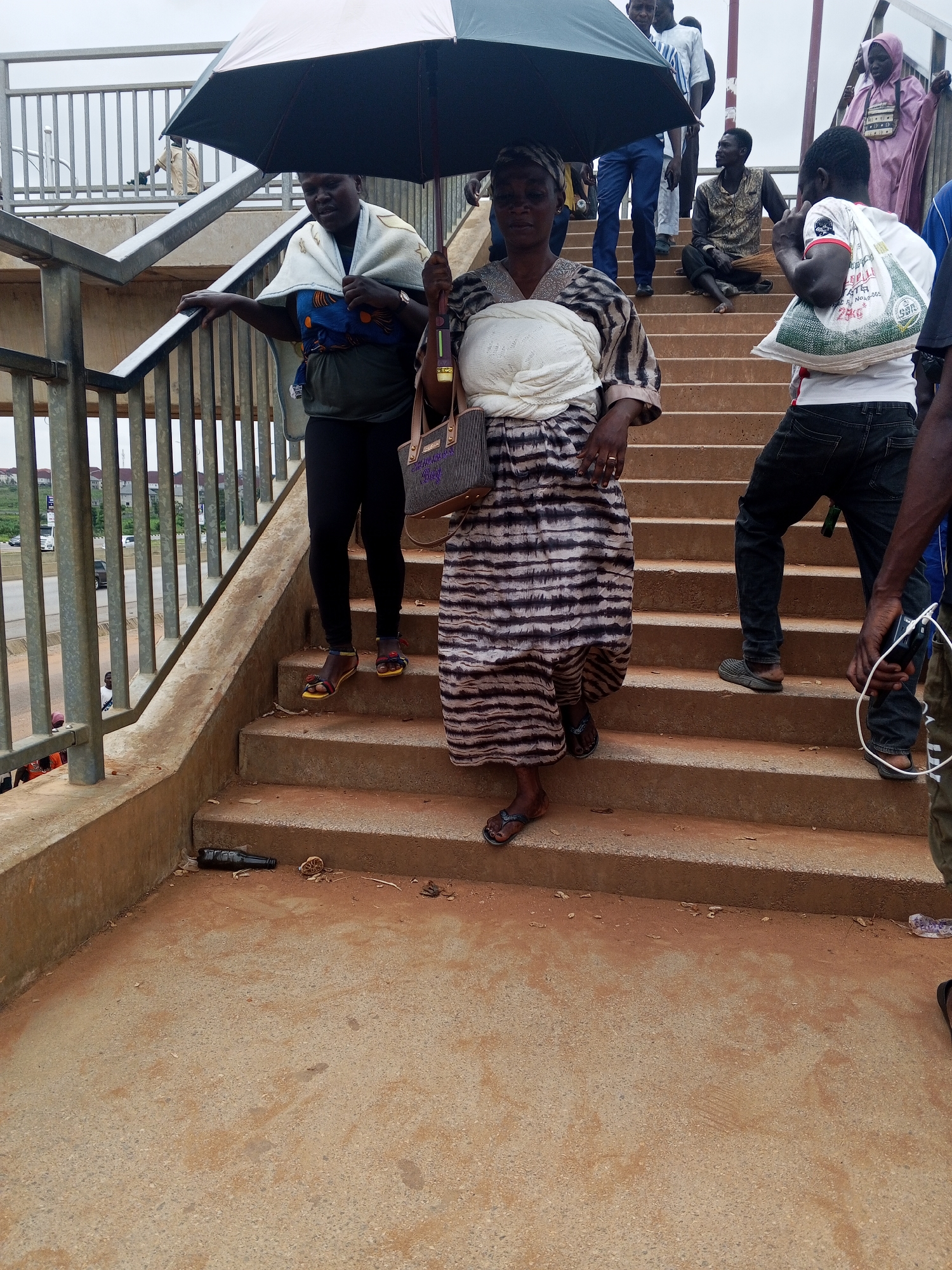 A group of people are on an outdoor pedestrian overpass in Sauka, Nigeria. The overpass has concrete stairs and metal railings. In the foreground, a woman is holding a black umbrella over herself and another person, who is wearing a white cloth around their shoulders. The woman is wearing a striped dress and dark leggings, and the other person is wearing a blue and green patterned garment. To the right, a man in a white t-shirt with writing on it is walking up the stairs, carrying a white bag. Behind him, another man in dark pants is also ascending. Further up the stairs, a man is sitting down with a broom, and a woman in a pink hijab is standing. A dark bottle and some debris are on the lower steps of the overpass. The sky is overcast.