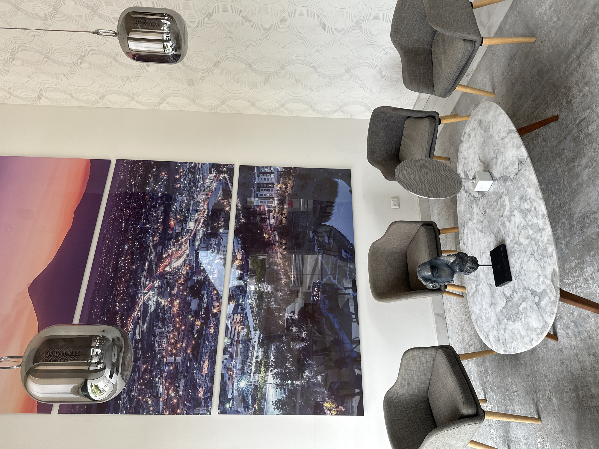 The image shows an interior design of what appears to be a waiting area or a modern living space. There is a sitting area with four gray upholstered chairs with wooden legs, arranged around a white marble oval table. On the table are decorations like a sculpture of a head and a modern cube object.
A set of three large framed pictures, each featuring a section of a city landscape, is on the left side of the image. The first shows a mountain at sunset, and the other two seem to show a city at night.
A modern, chrome pendant lamp is seen hanging from the ceiling. The ceiling has a light-colored wallpaper with a subtle wave pattern.
Given the cityscape in the pictures, this image could potentially depict a location in Puebla, Mexico, if the skyline and urban layout depicted in the art align with the actual city's appearance. However, without additional information or a more specific viewpoint, it is hard to confirm definitively. The overall atmosphere is modern and stylish, with an emphasis on contemporary art and furniture.