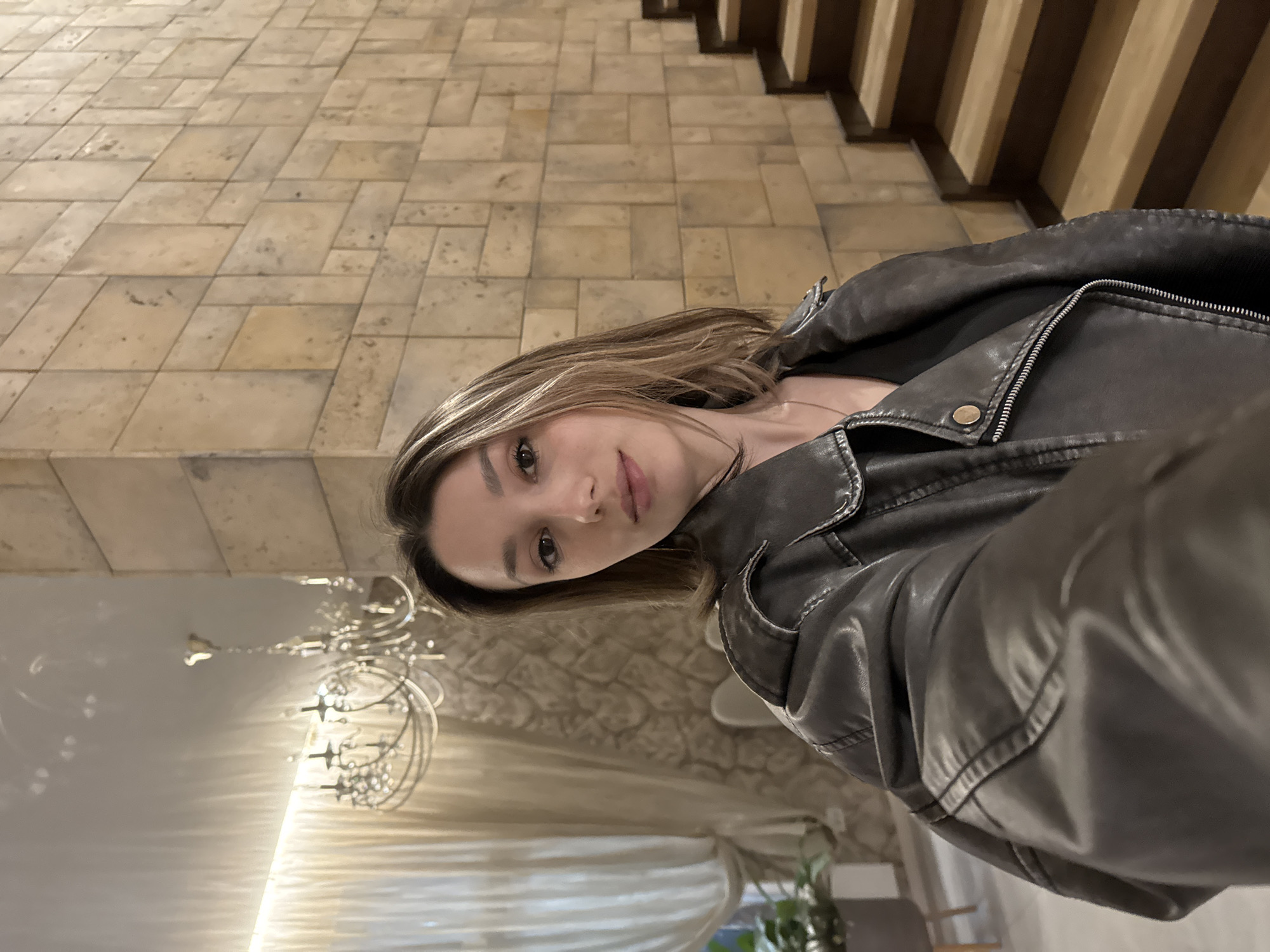The image is a selfie of a woman in an indoor setting. She is wearing a grey leather jacket over a dark shirt, and her hair is light brown with blonde highlights. Her facial expression is neutral.
Behind the woman is a wall covered in tan, rectangular tiles, possibly made of stone. To the right of the wall are wooden stairs with dark brown treads and lighter risers. In the lower left corner of the image, there is a white ceiling with a large, ornate chandelier hanging from it. There is also a window with sheer white curtains, and a potted plant can be seen near the window.