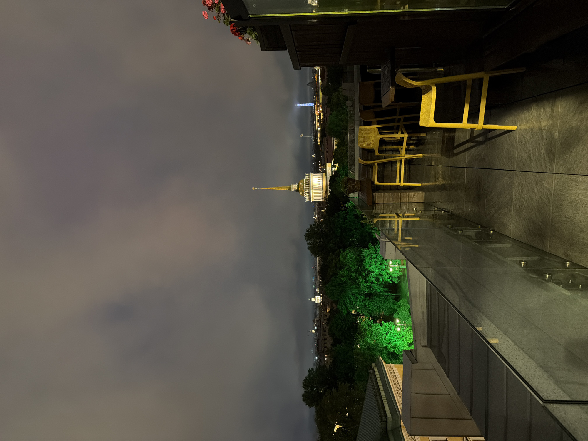 This is a nighttime, horizontal, medium shot taken from a rooftop, looking out over a city skyline. The foreground features a glass-paneled railing and a wet stone patio with several bright yellow bar stools and tables. In the middle ground, a cluster of trees is illuminated with bright green lights. The background displays a city skyline under a cloudy, overcast sky. Prominent structures include a tall, golden-domed building with a spire and a distant tower that is illuminated with blue light. The scene appears to be set in St. Petersburg, Russia, with the golden-domed building likely being the Admiralty Building and the blue-lit tower potentially being the Peter and Paul Fortress or a similar landmark.