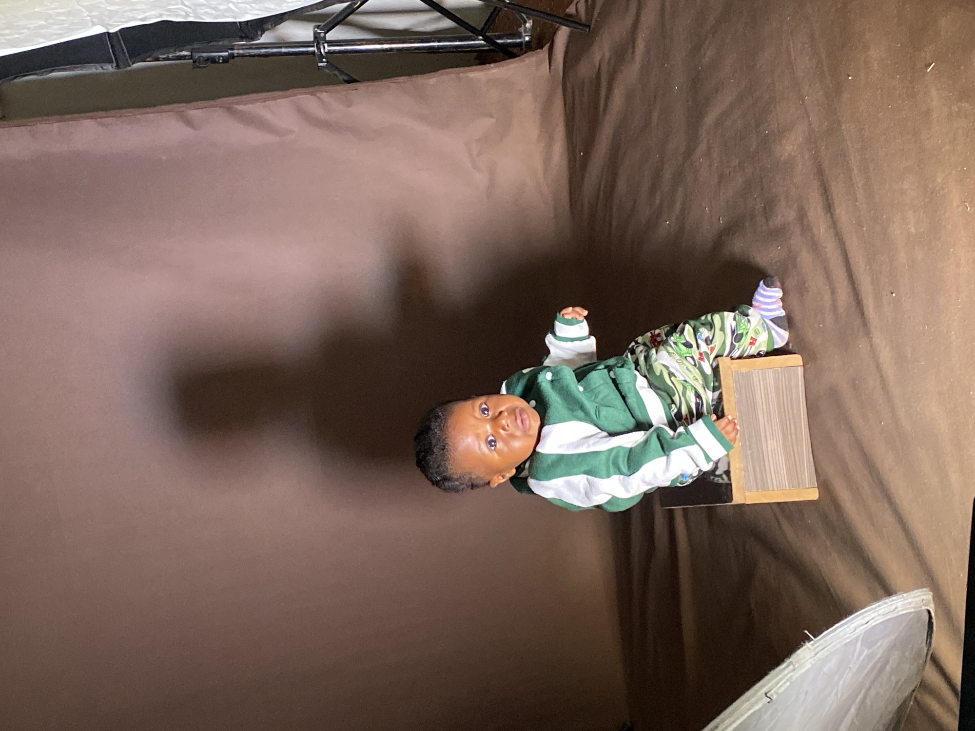 A baby is the central subject of this photograph, positioned in what appears to be a makeshift indoor photography studio. The baby, likely a toddler, is dressed in a green and white striped long-sleeved top, camouflage-patterned trousers, and mismatched striped socks – one blue and white, the other a lighter color. The baby looks directly at the camera with a wide-eyed, curious expression.
The baby is either sitting or standing on a small, light-colored wooden stool or box that has a textured top surface. The background is a plain, dark brown fabric, likely a photography backdrop, which is draped and folded, creating an enclosed setting. Visible at the top left of the frame are parts of a metal stand or frame, indicative of photography equipment. In the bottom right corner, the edge of a silver and white reflector or softbox can be seen, suggesting controlled lighting for the shoot.
The overall scene depicts a professional or semi-professional baby photoshoot. The specific location is not visually discernible from the image itself, but the provided context indicates Vom, Nigeria. There is no visible text within the image.