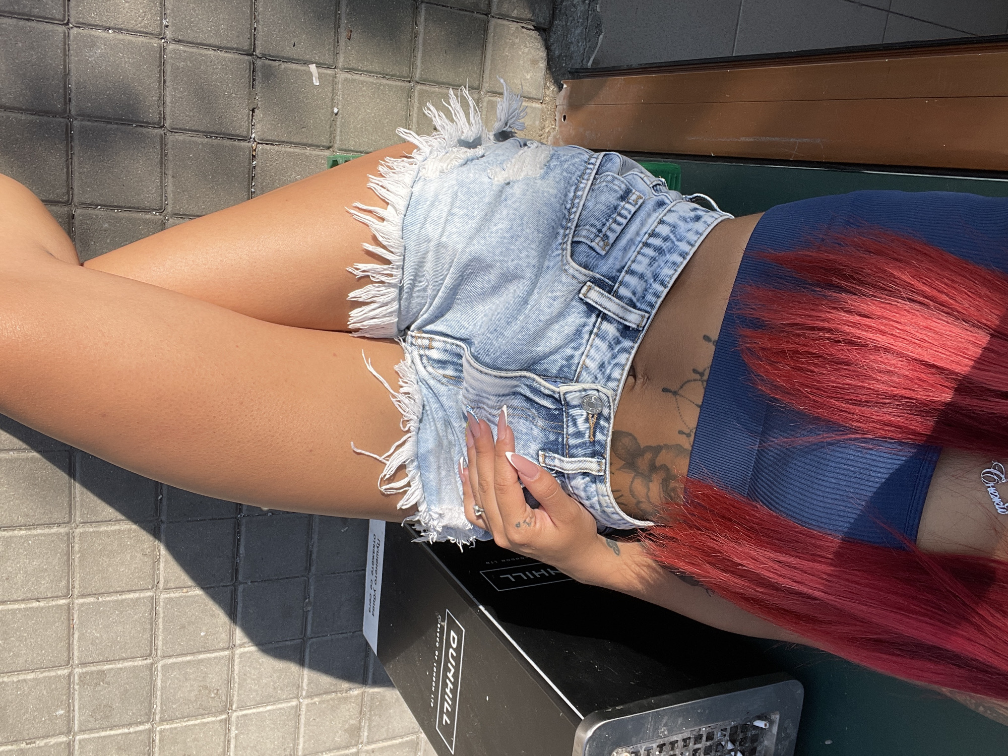 The image shows a close-up of a woman's torso and legs, from just below her chest to her mid-thighs. She is wearing a dark blue ribbed crop top and light-wash denim shorts with frayed hems. The woman has bright red hair that falls over her shoulders. A tattoo is visible on her abdomen. Her right hand, with manicured nails, rests on her hip. To her right, partially visible, is a tall, black trash receptacle with a metallic grate on top and branding that reads "DUNHILL" and "CIGARETTES OF LONDON LTD." Below the Dunhill branding, there is text in Bulgarian that appears to be a warning related to smoking. The background consists of a tiled sidewalk and a portion of a building with a dark green and brown facade. The scene is illuminated by bright sunlight, casting shadows on the ground. The overall setting suggests an outdoor urban environment, possibly near a commercial establishment. The location is identified as Sofia, Bulgaria.