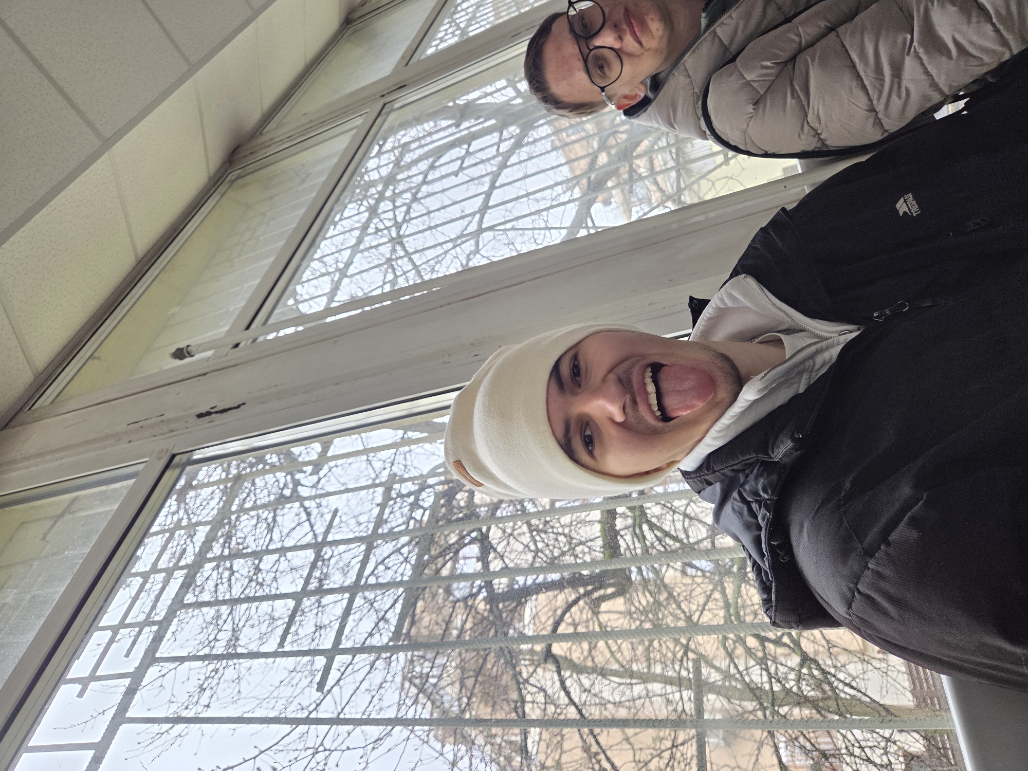 Two young people are taking a selfie, looking down at the camera. One is wearing a cream-colored beanie and a black jacket and has their tongue sticking out. The other is wearing glasses and a tan puffer jacket. They are standing in front of a window with a metal grate. Through the window, bare tree branches can be seen. The location appears to be inside a building, possibly near a window overlooking an urban area, based on the trees and general building structure visible through the window, which may be located in Sofia, Bulgaria.