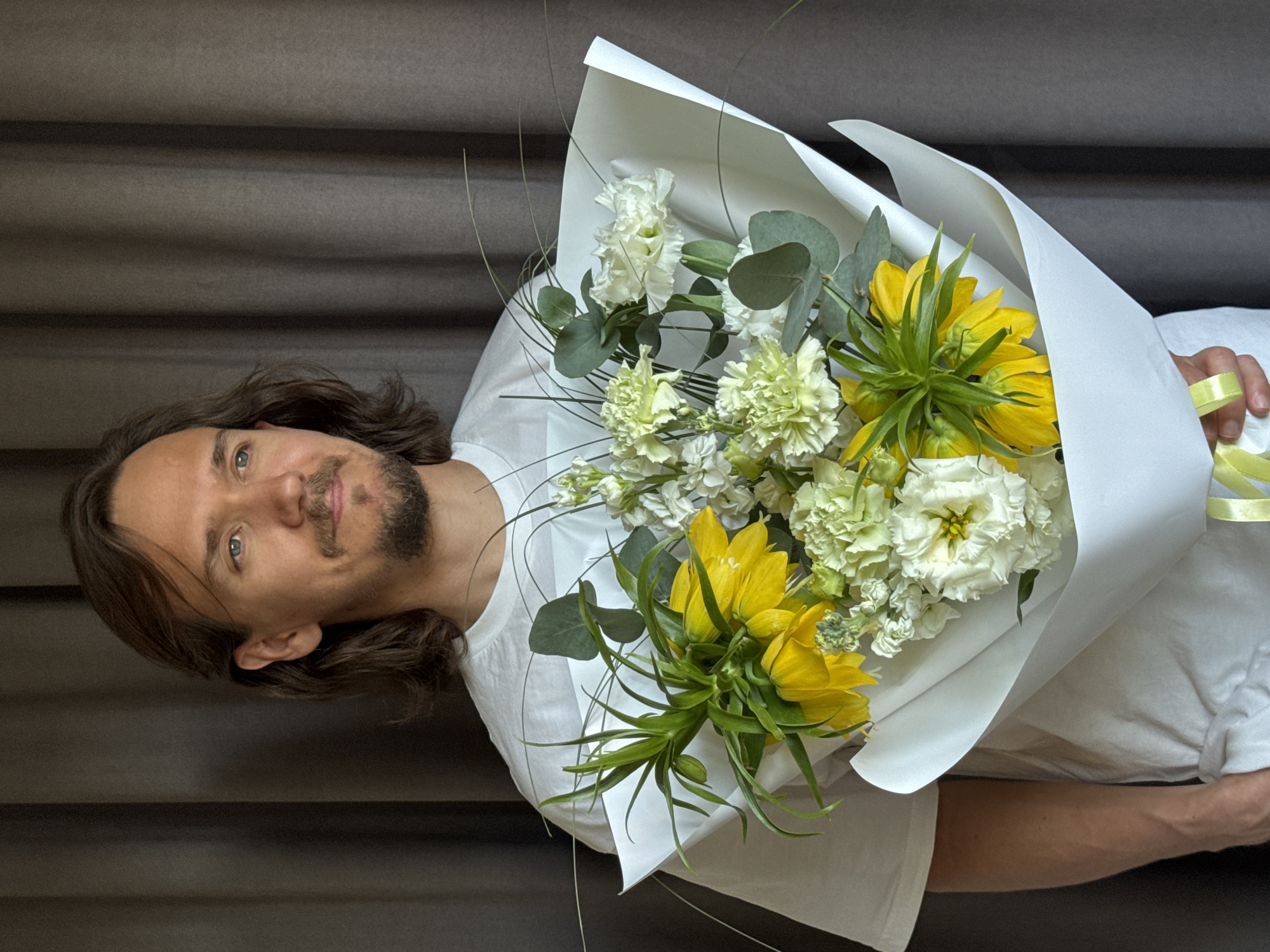 A man with long brown hair, a light beard, and a white T-shirt is holding a bouquet of flowers. The bouquet consists of white and yellow flowers wrapped in white paper with a light yellow ribbon. The background is a gray, textured wall. The location is unspecified and cannot be determined to be in Izhevsk, Russia.