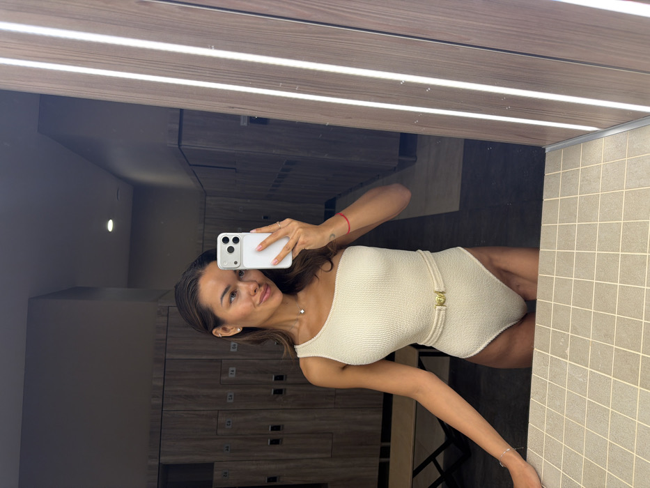 A single adult female is depicted taking a mirror selfie in a locker room located in Krasnoyarsk, Russia. The subject has brown hair, which appears wet or styled back, and is wearing a cream-colored, textured, one-shoulder one-piece swimsuit. A thin, woven, light-colored belt with a gold-toned buckle encircles her waist. She wears a delicate necklace with a small pendant, stud earrings, and a red string bracelet on her right wrist; a faint tattoo is visible on her right inner forearm. She holds a white smartphone with a multi-lens camera array in her right hand, pointed towards her reflection. Her left forearm rests on a light grey tiled counter in the foreground. The background, reflected in the mirror, displays rows of modern, light wood-effect lockers with black handles, some numbered (e.g., C1, C3, C4, C5, C6, C7, T.A., T.B.). A wooden bench is visible to the left within the reflection. The area is illuminated by overhead recessed lighting and vertical light strips integrated into the right side of the mirror. No other individuals or significant activities are present in the visible scene.