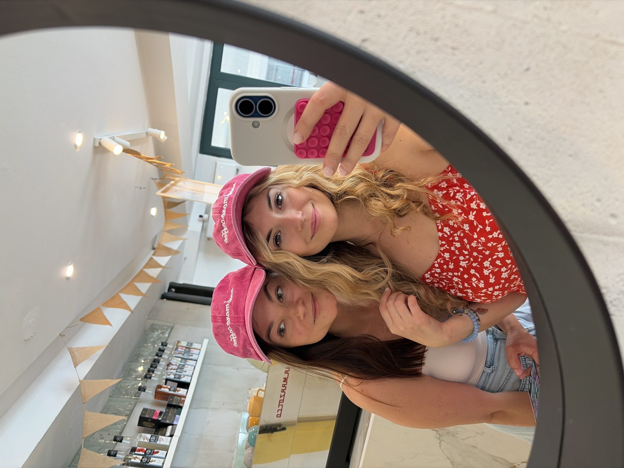 Two young women are reflected in a round mirror. Both are wearing pink baseball caps. The woman in the foreground has dark brown hair and is wearing a white tank top and light blue denim shorts. The woman in the background has blonde curly hair and is wearing a red floral-print top. She is holding a smartphone with a white case and a pink pop-socket. The mirror reflects the interior of a shop, which appears to be a coffee shop or boutique. There are shelves with products, decorative flags hanging from the ceiling, and what appears to be a coffee machine. The location is likely in Budva, Montenegro, based on the information available in the image and its description.