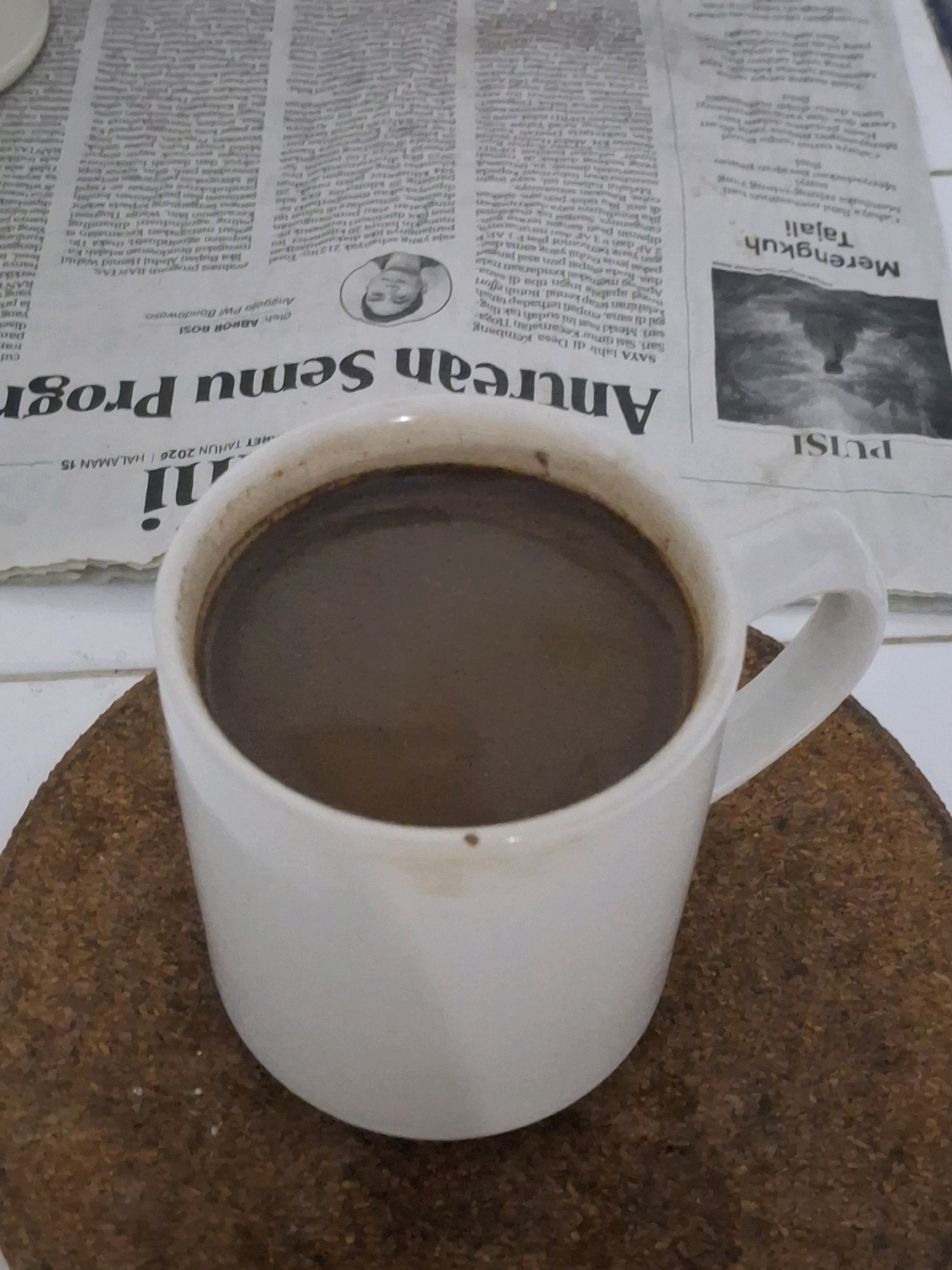 A white mug of dark liquid, likely coffee, sits on a cork coaster atop a newspaper. The newspaper has visible text and images. The text is in Indonesian. One headline visible reads "Anurean Semu Proger". Another section has text in a smaller font, and a headline that reads "Merengkuh Tajali". A small, black and white portrait of a man is present on the newspaper. The overall scene is likely indoors, given the newspaper and the presence of a mug. The environment is uncluttered and suggests a moment of quietude. The presence of Indonesian language text on the newspaper and the origin of the city name "Lumajang" in Indonesia strongly suggests the image was taken in Indonesia.