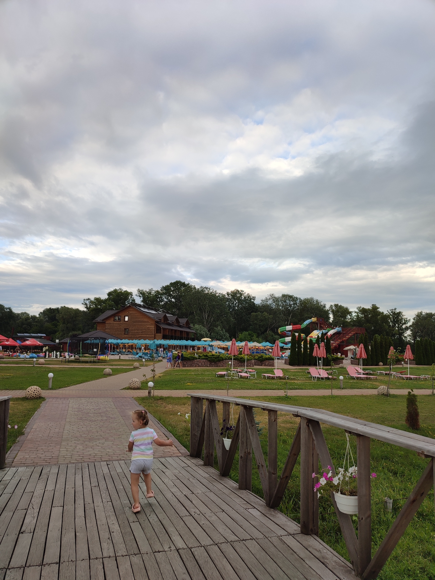 This media file captures a scene at a water park on an overcast day, likely in the late afternoon or early evening. In the foreground, a young girl with blonde hair pulled into pigtails is walking across a wooden bridge. She is wearing a striped t-shirt and shorts. The bridge has wooden railings with decorative wooden supports and hanging flower baskets.
Beyond the bridge, the water park unfolds. A pathway leads towards a large wooden building with a dark roof. To the right of the building, colorful water slides are visible, indicating a recreational setting. Numerous pink beach umbrellas are dotted across a well-maintained lawn, with lounge chairs placed beneath them, suggesting areas for relaxation. Some trees and dense foliage line the background, framing the park. The sky is filled with heavy, grey clouds, creating a subdued lighting condition. The overall atmosphere appears calm and relaxed, with no explicit indication of intense activity, though the presence of the water park and lounge areas suggests it is a place for leisure. The location is contextually identified as Shcherbani, Ukraine.