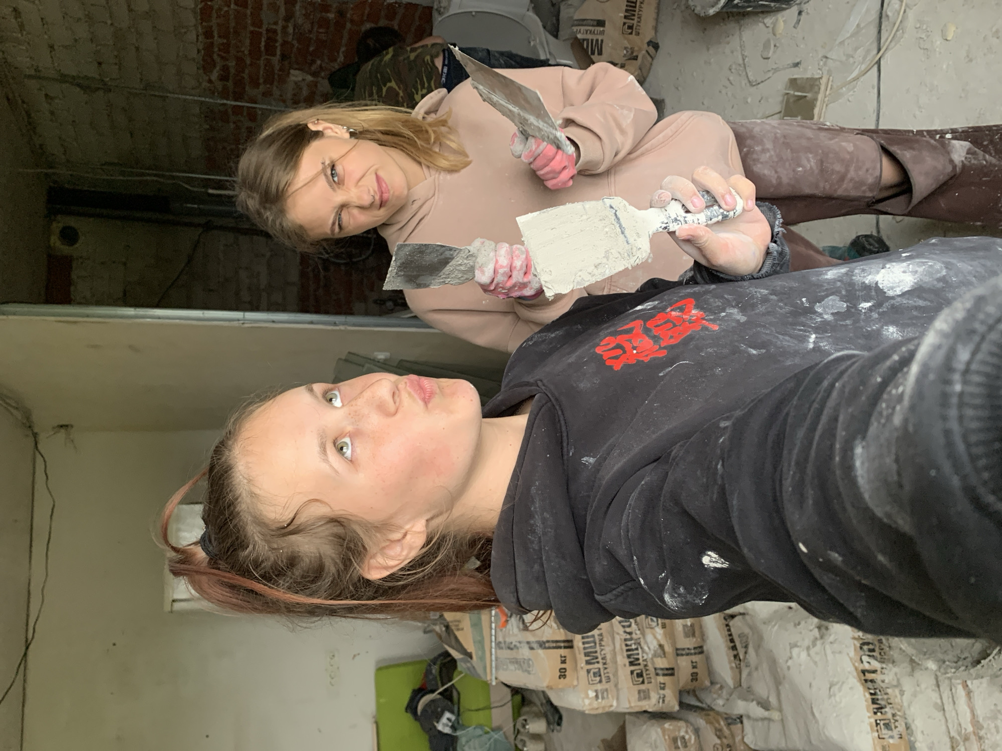 Two young women are posing for a selfie in what appears to be a construction or renovation site. The foreground shows a young woman with light skin, green eyes, and light brown hair with some pinkish-red streaks. She is wearing a black hoodie with some red lettering or design on the chest and is splattered with white material, likely plaster or paint. She is holding a trowel with white plaster or compound on it. Behind her is another young woman with similar complexion and blondish-brown hair. She is wearing a light pink hoodie, dark brown trousers, and pink gloves and is also holding a trowel. Bags of what appear to be cement or plaster mix are stacked in the background. The location is Фоки, Russia.